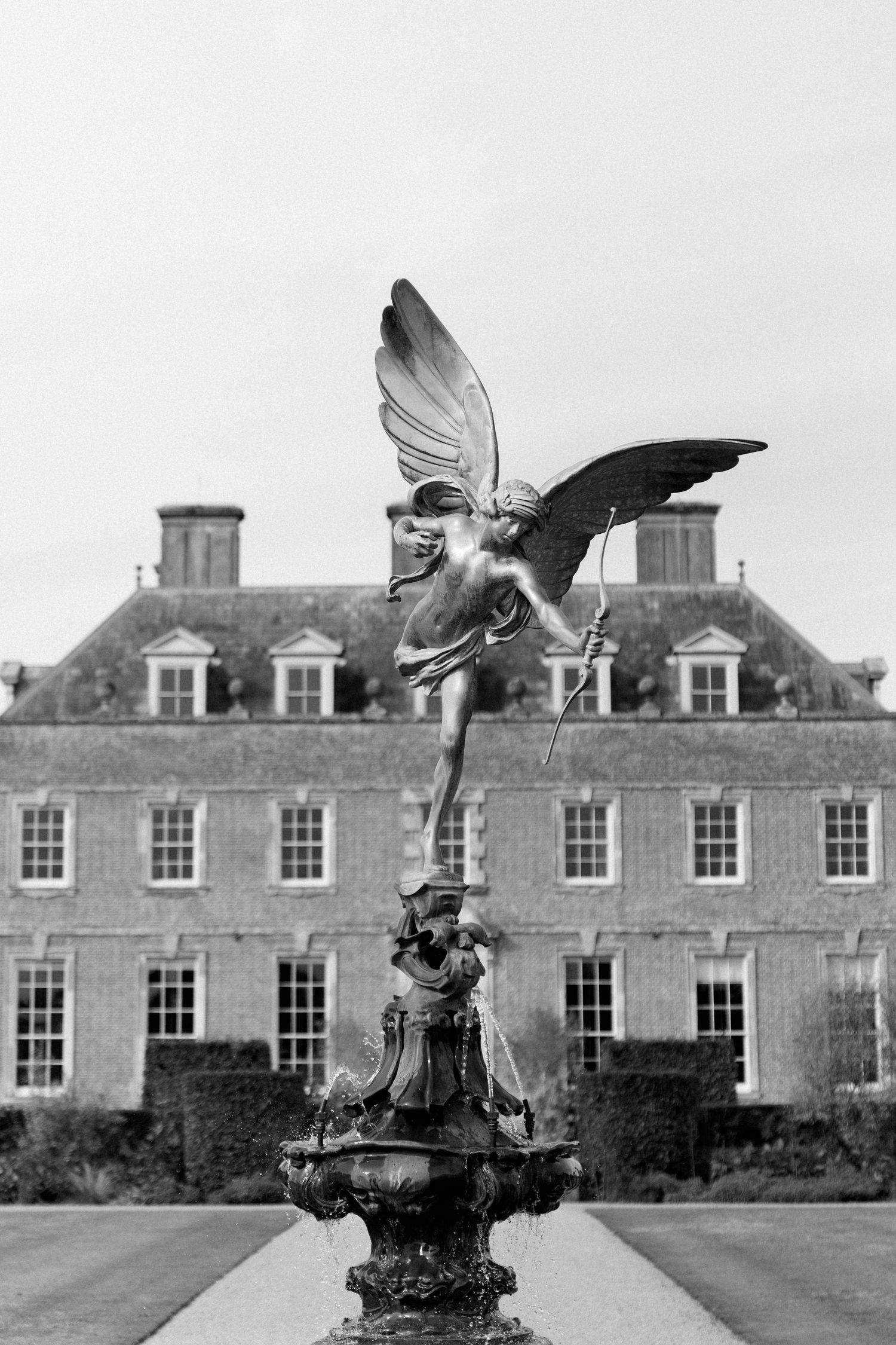 Black and white photo of an angel statue fountain with grand Georgian manor house in background.