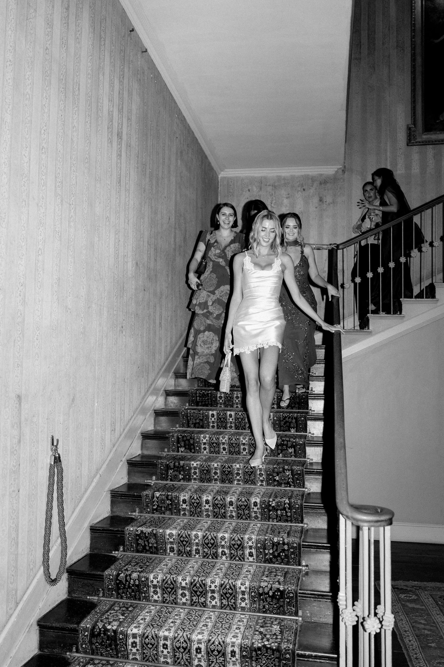 Bride in white mini dress leads bridesmaids down a patterned staircase at a wedding venue.