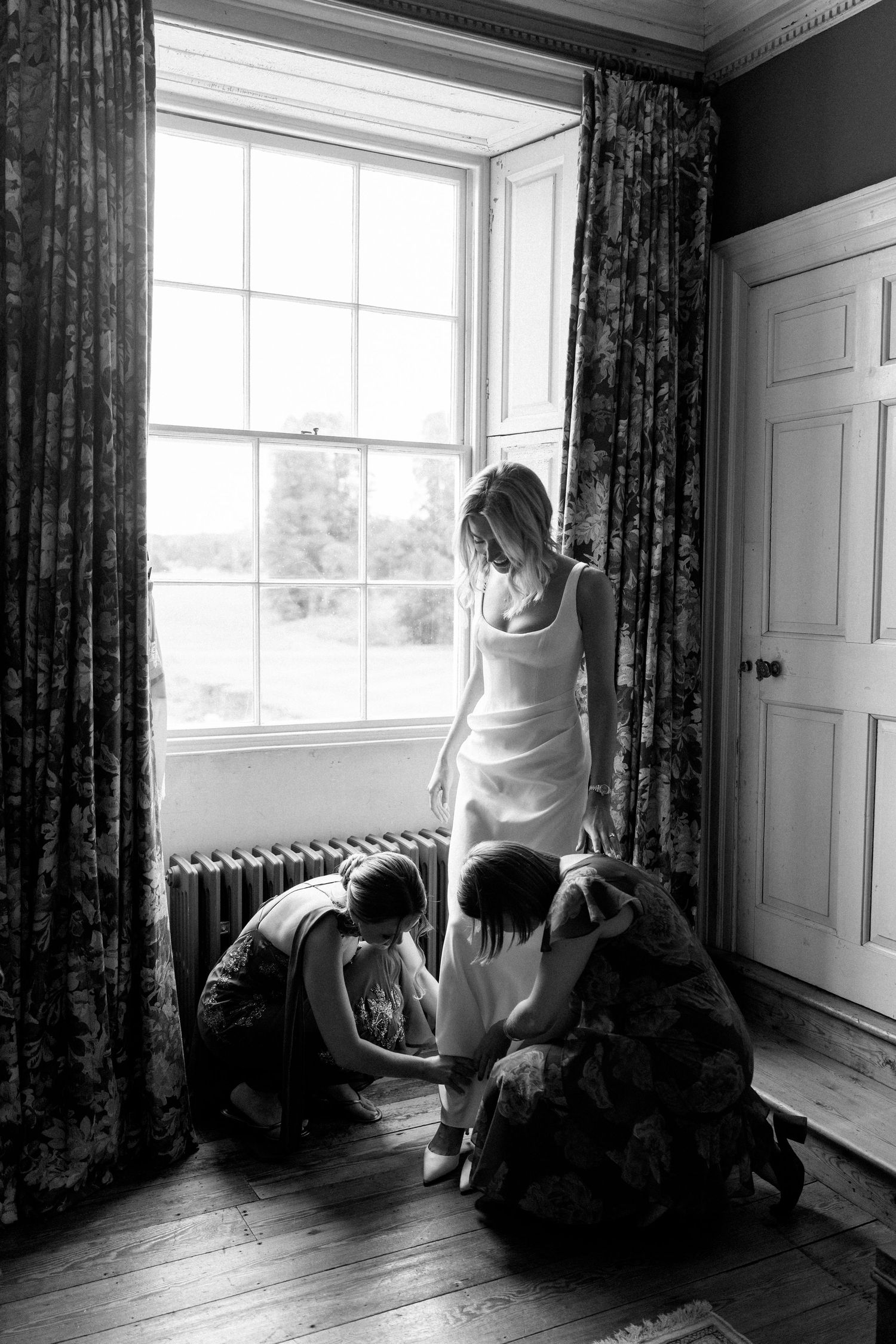 Black and white photo of bride being helped into shoes by bridesmaids near a large window.
