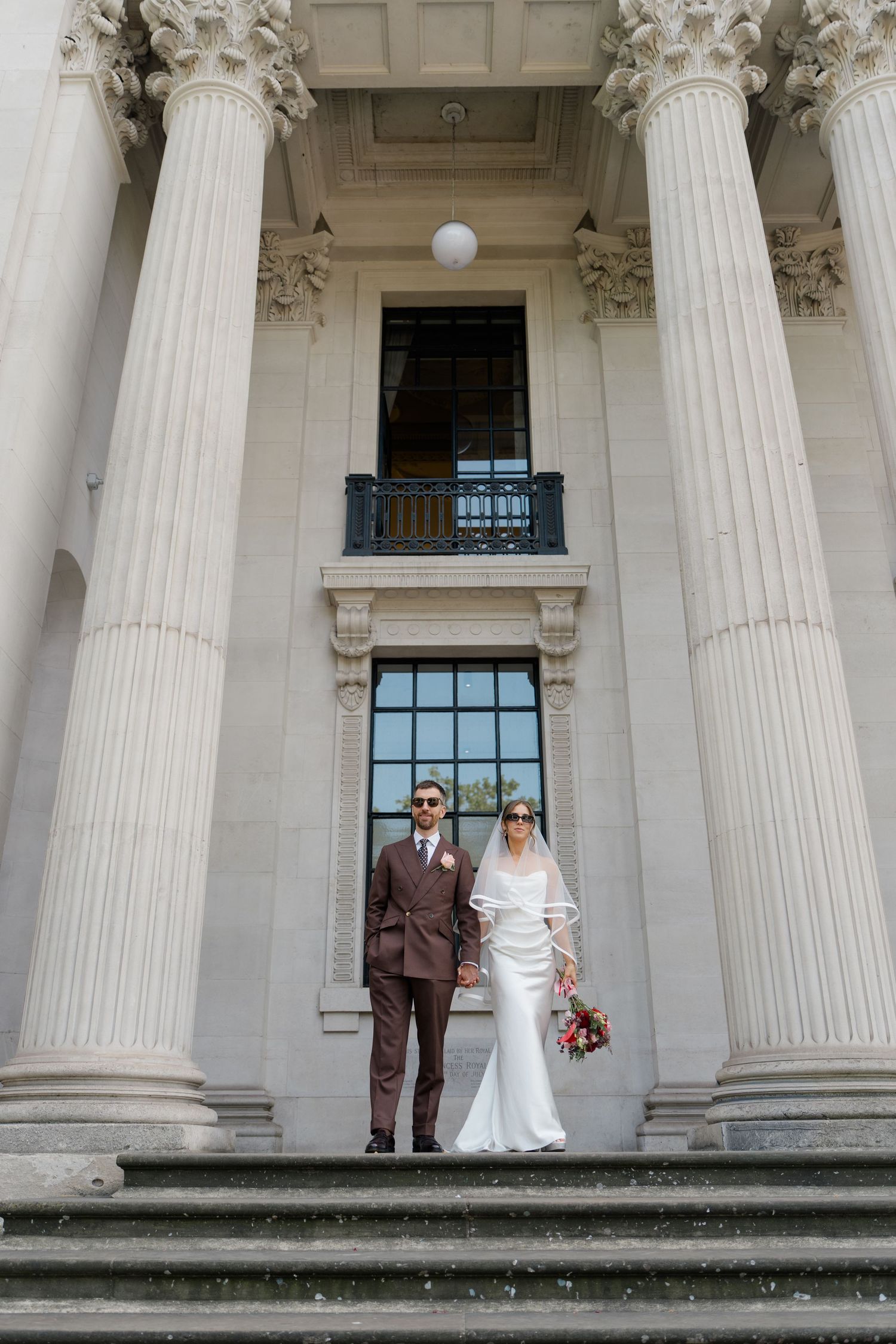 Bride and groom pose on steps between towering classical columns at an elegant wedding venue exterior.