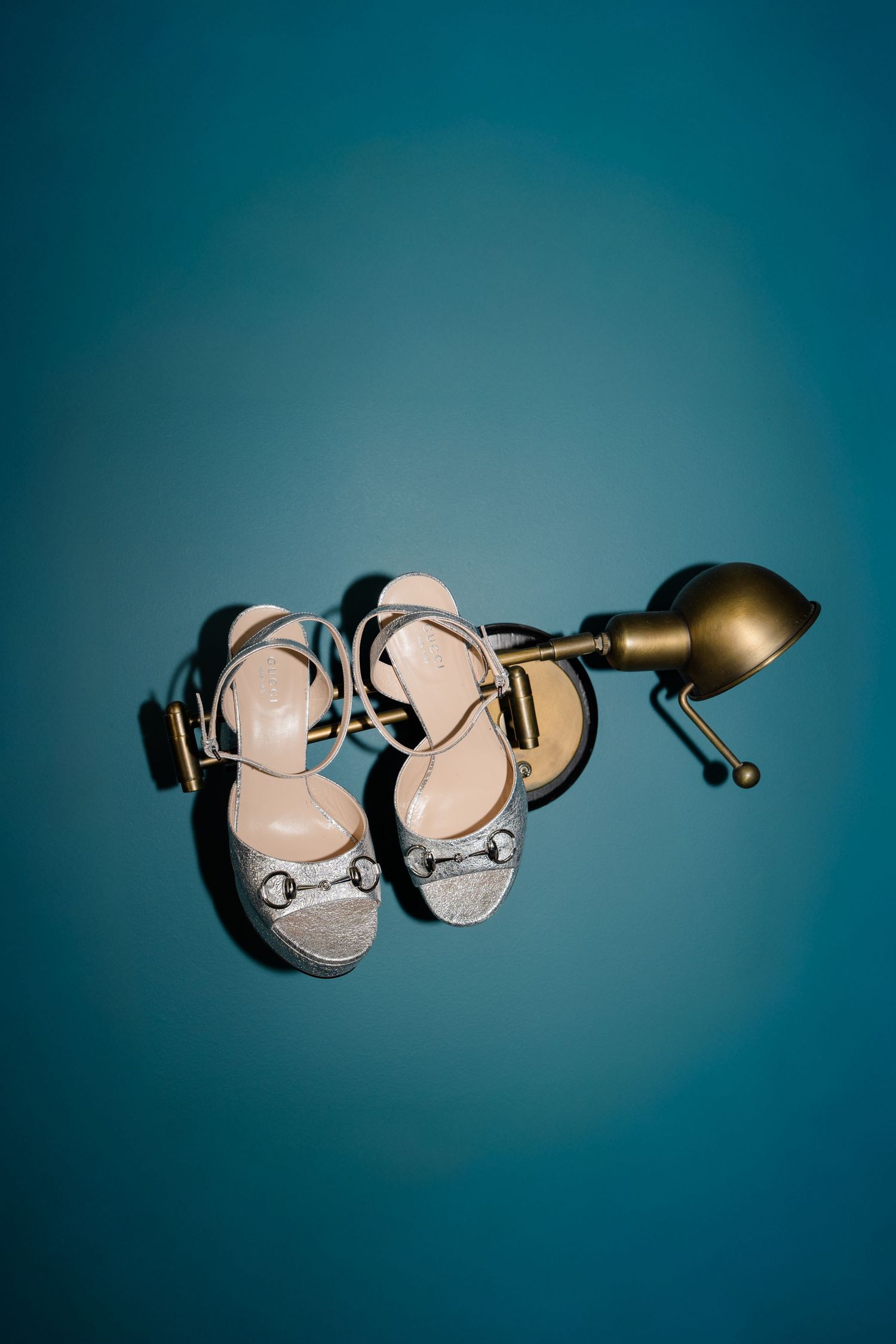 Silver glitter wedding heels with gold horsebit detail resting on a brass lamp against a teal background.