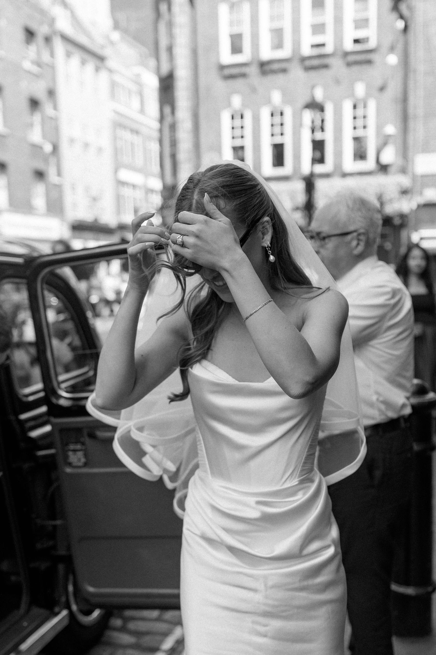 Bride in white satin gown adjusts sunglasses beside black London taxi cab, black and white photo.