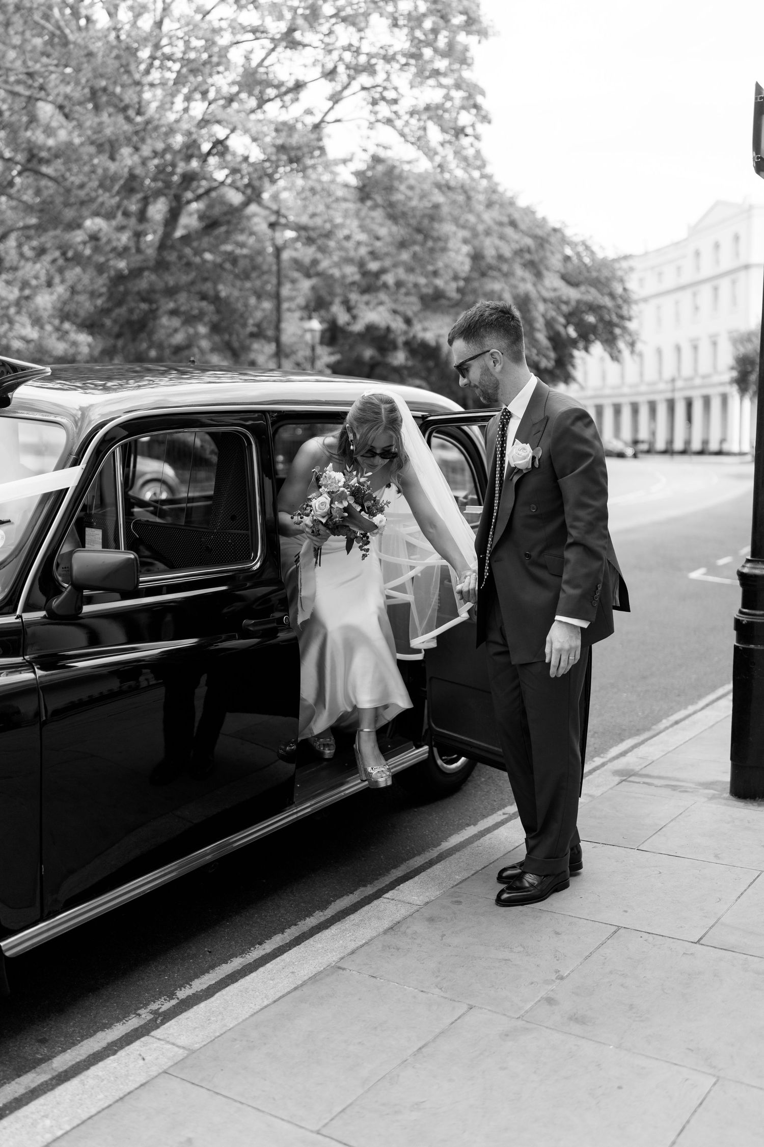 Groom helping bride exit a classic black London taxi cab on a city street in black and white.