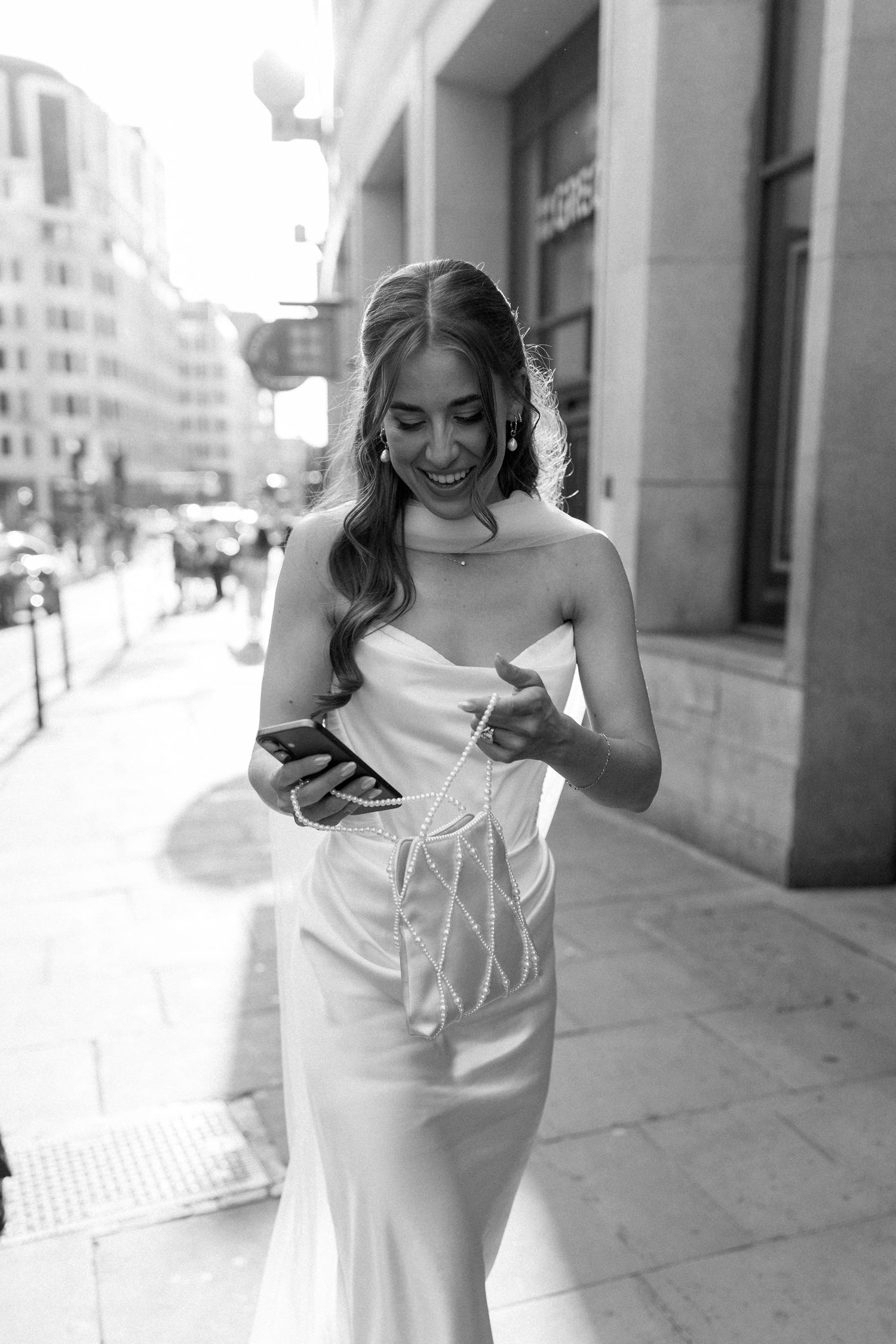 Smiling bride in strapless white gown checking phone on urban city sidewalk in black and white photo.