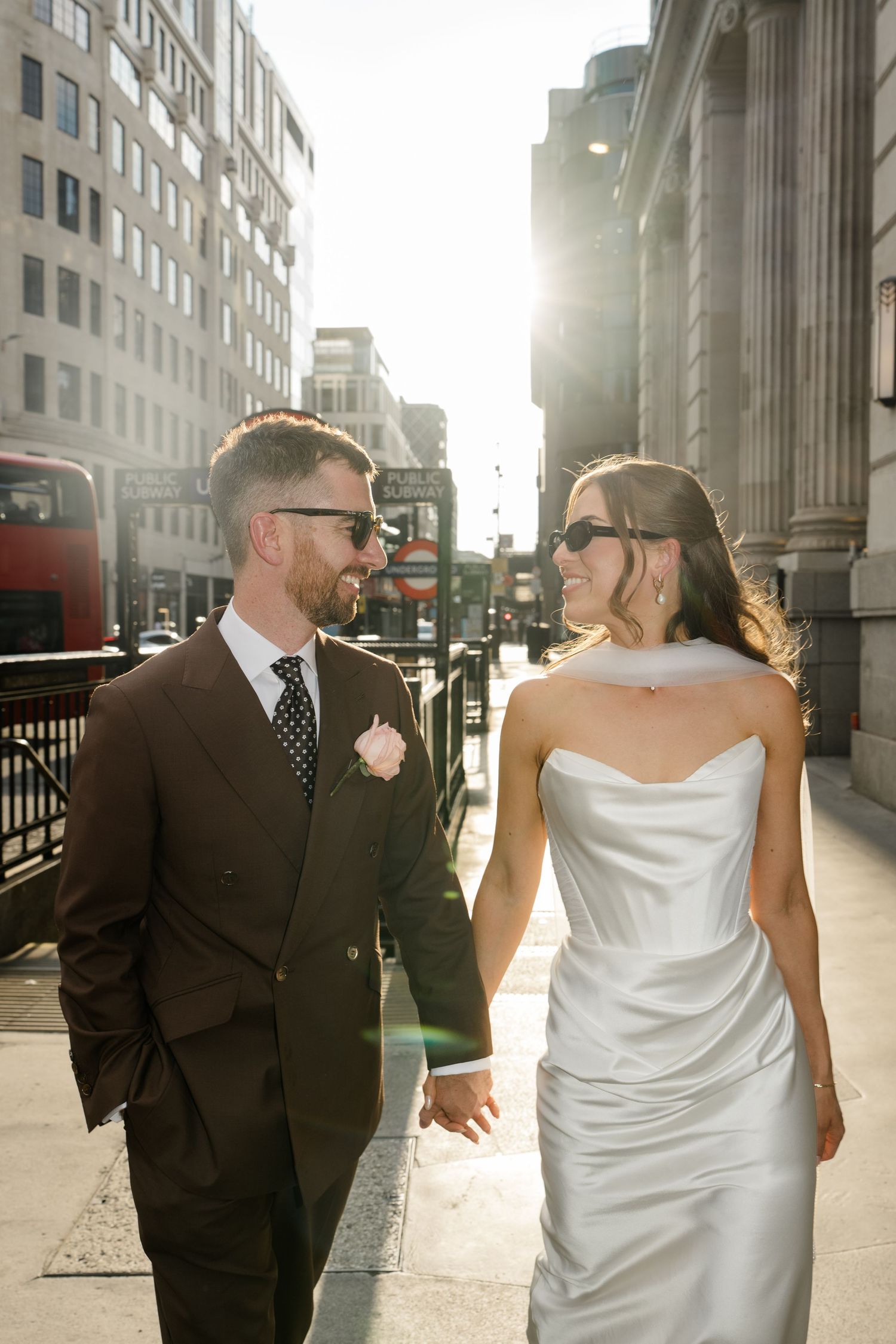 Newlyweds holding hands walking through sunny London streets, bride in white satin gown and groom in brown suit.