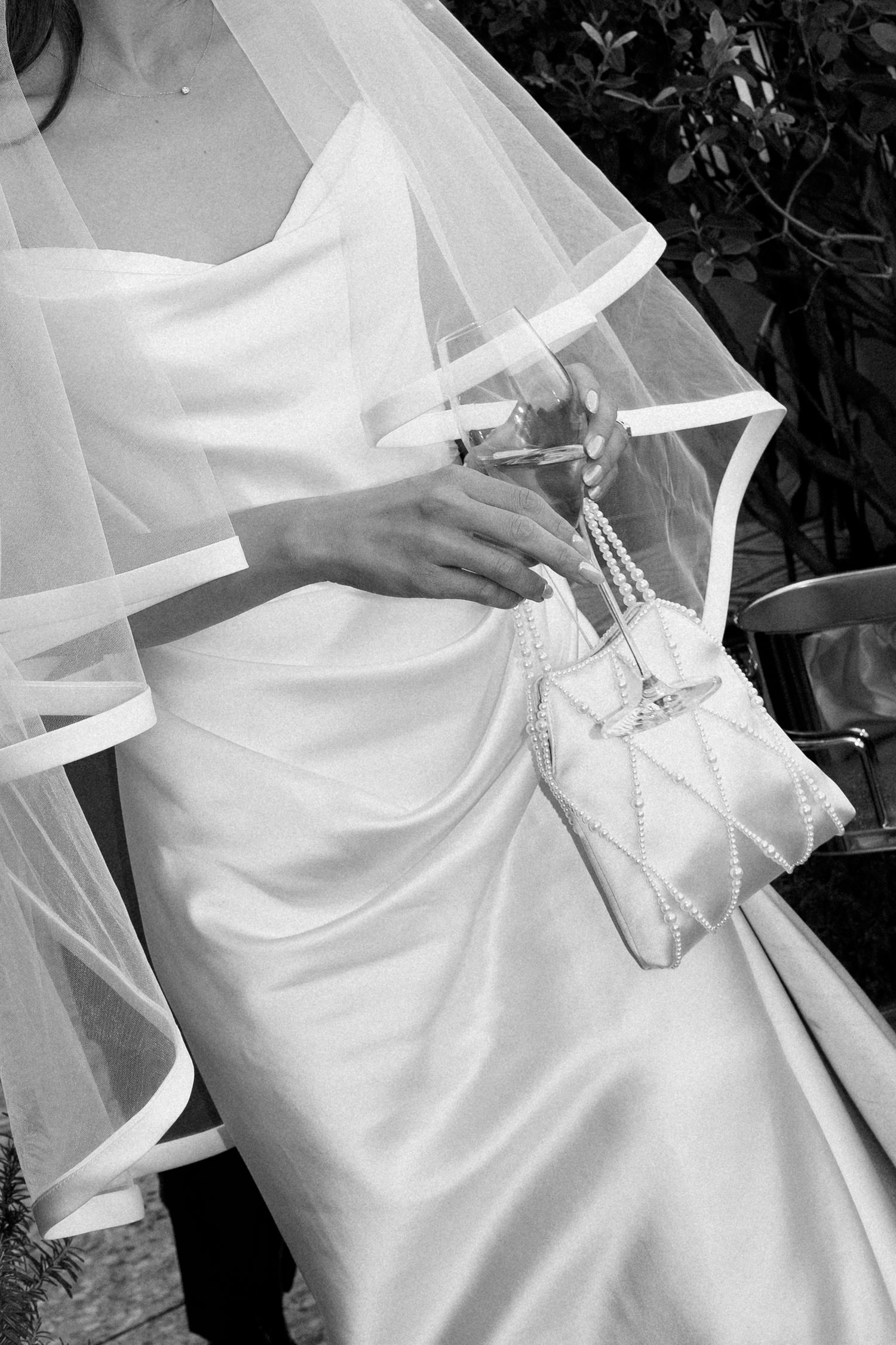 Close-up of bride in satin gown holding pearl handbag and champagne glass beneath flowing white wedding veil.