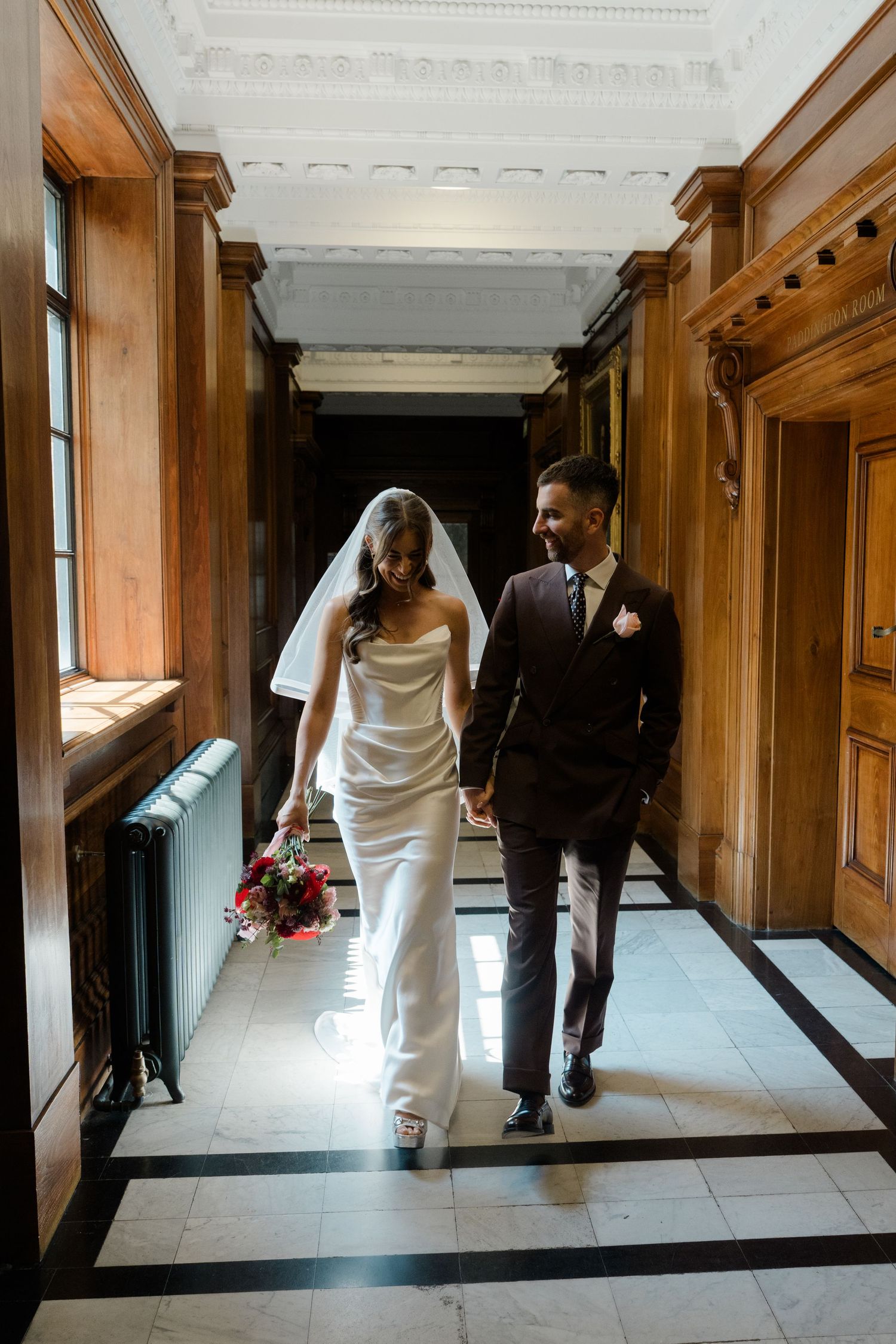 Bride and groom walk hand in hand through an elegant wood-paneled hall on their wedding day.