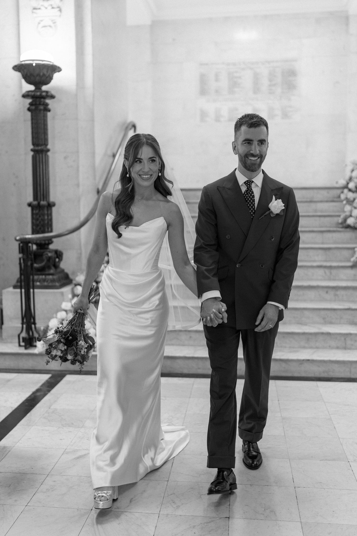 Smiling bride and groom holding hands walking down grand marble staircase in black and white photo.