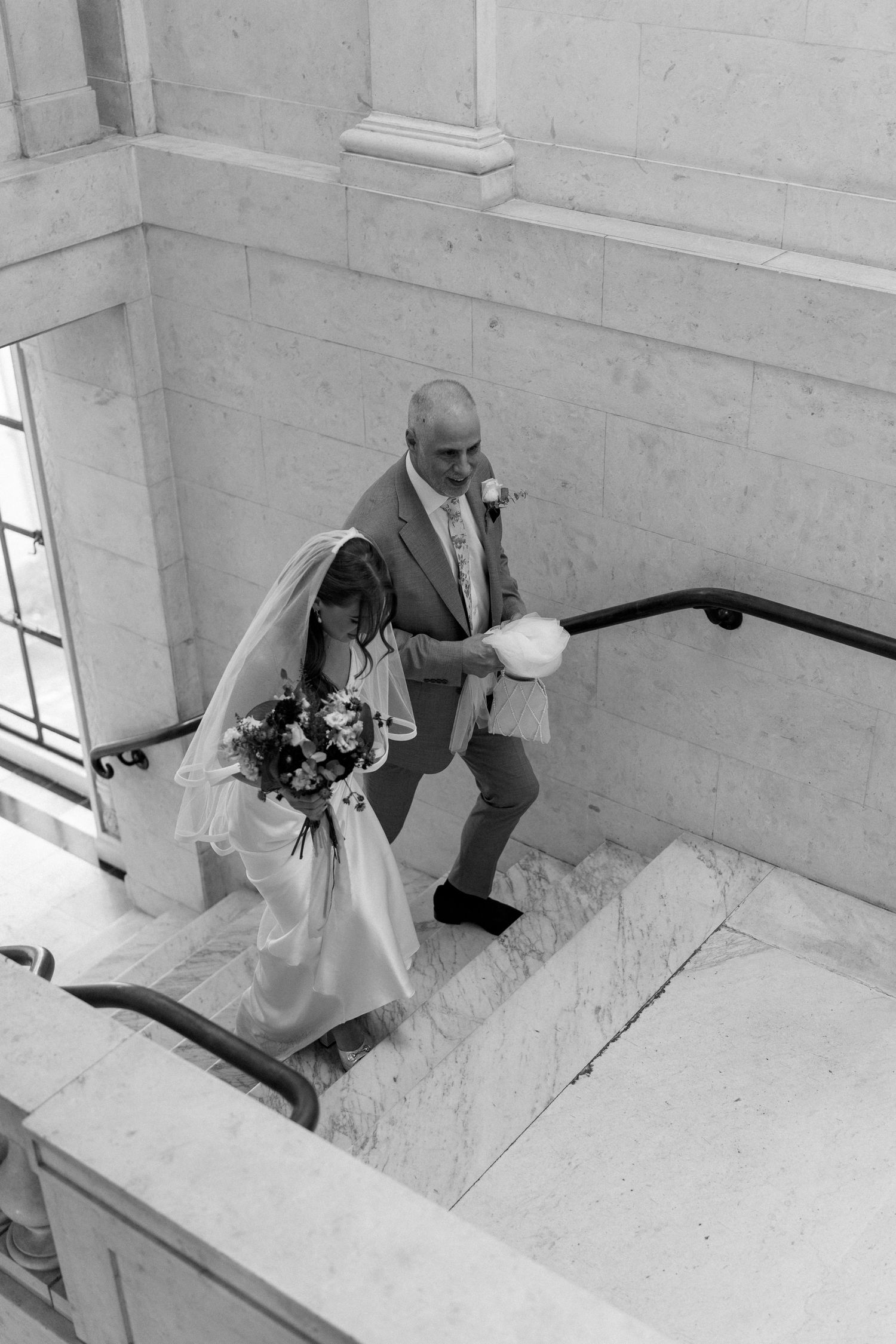 Aerial black and white photo of bride and groom ascending marble staircase with bouquet.