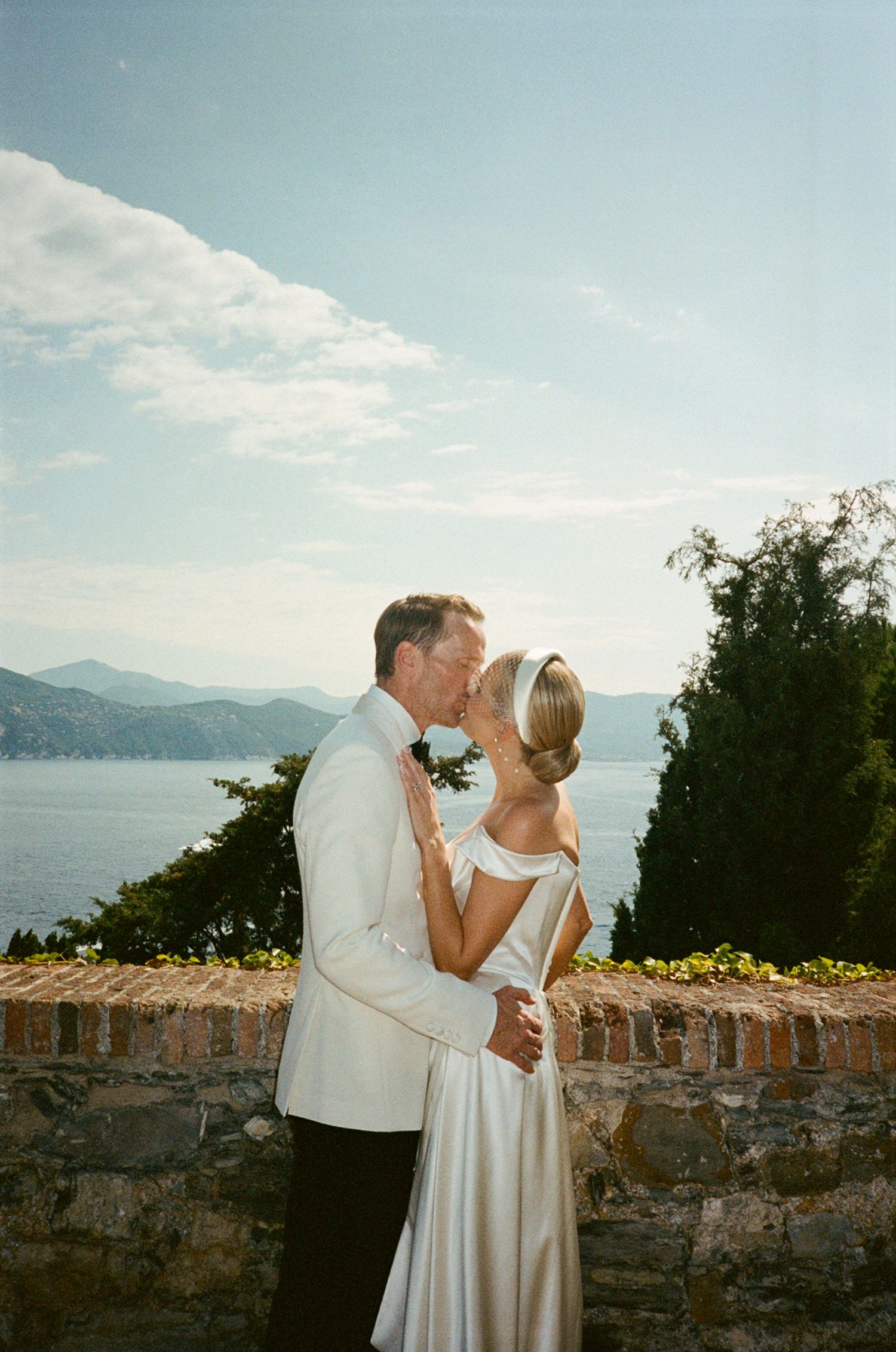 Bride and groom kissing at outdoor wedding venue overlooking blue sea and mountains in white tuxedo and satin gown.
