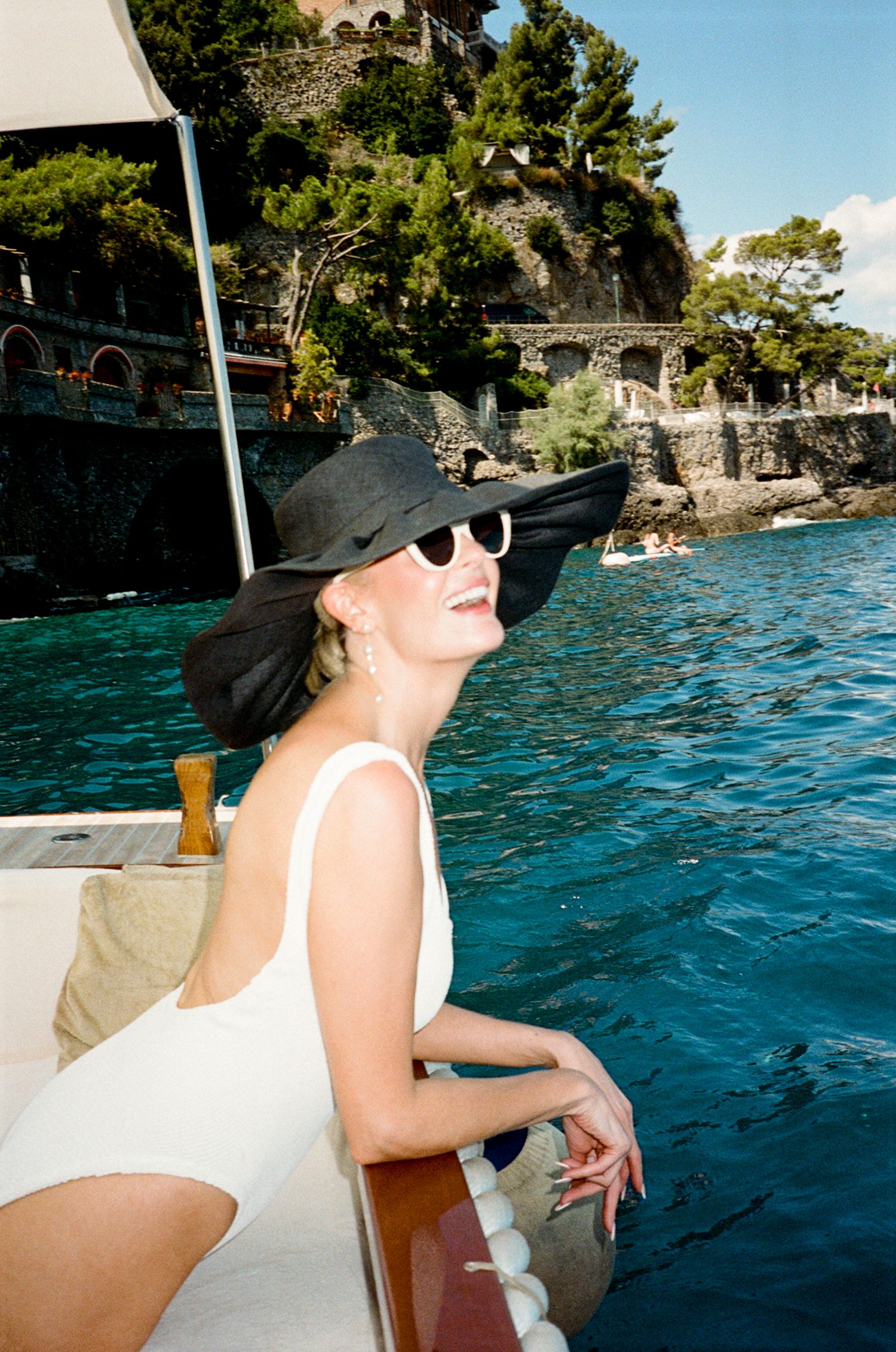 Woman in white swimsuit and black wide-brim hat enjoying a scenic boat ride on turquoise waters.