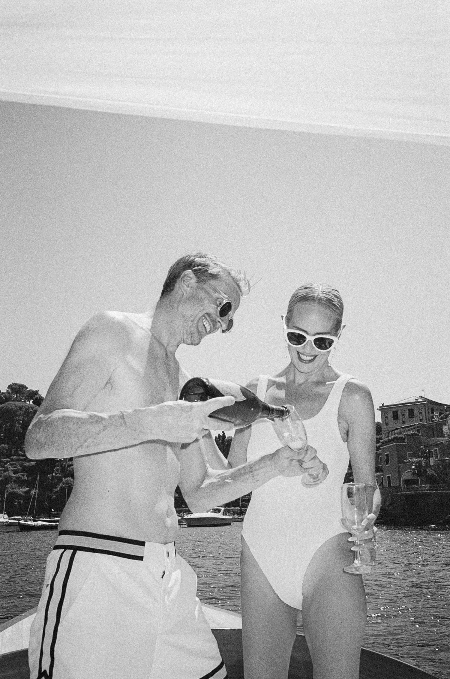 Black and white photo of couple in swimwear sharing champagne and laughing on sunny boat in Italy.