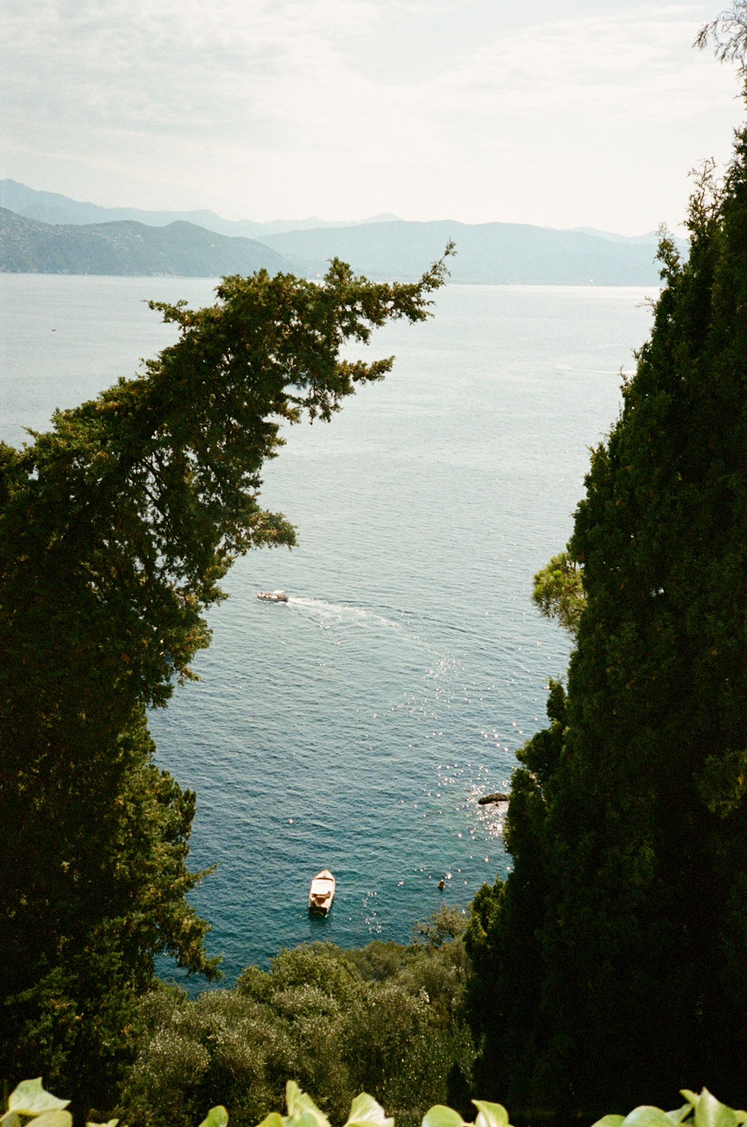 Aerial view of blue Mediterranean sea framed by lush green trees with boats on water and mountains in background.