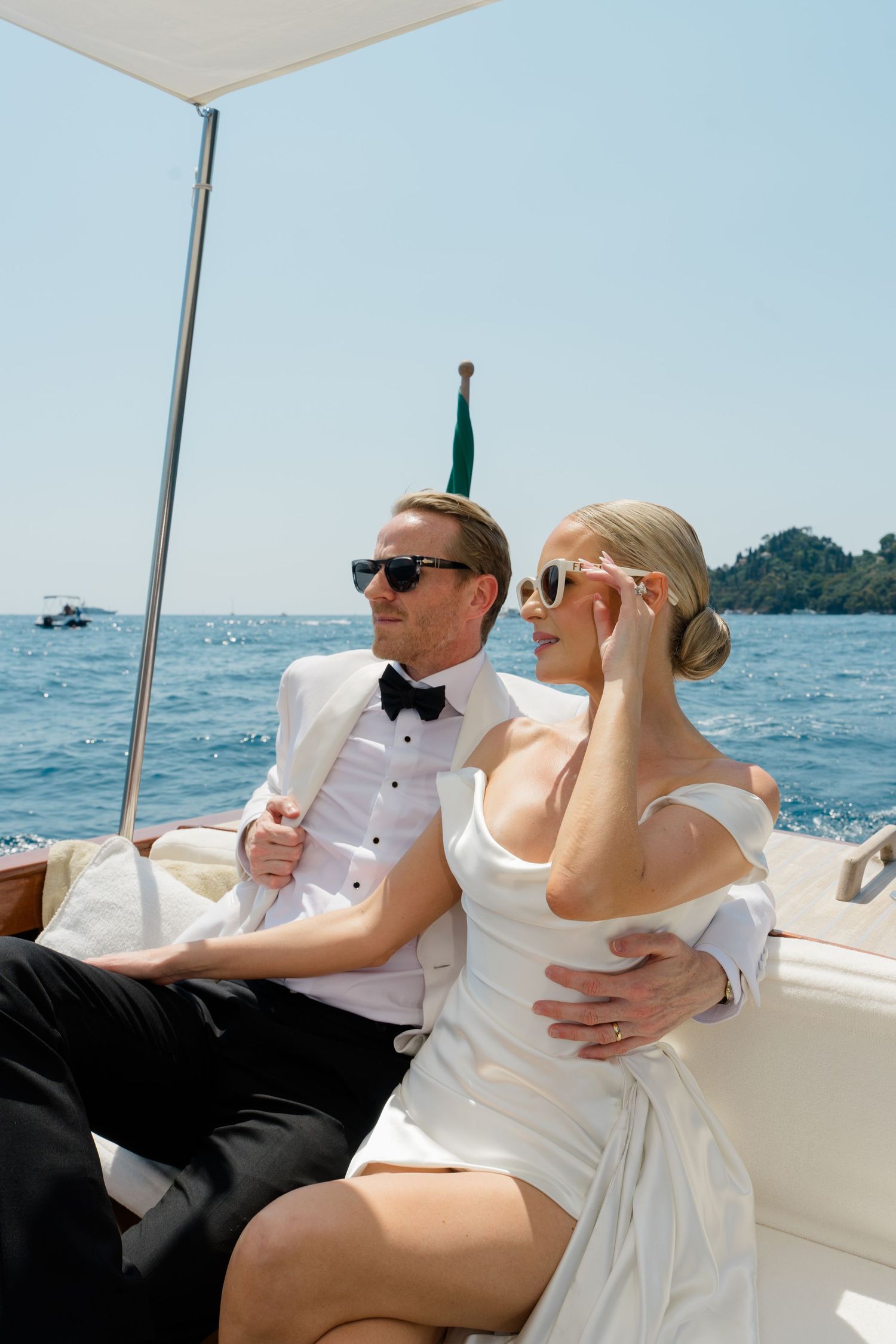 Elegant bride and groom in white attire and sunglasses relaxing on a luxury yacht on a sunny day.