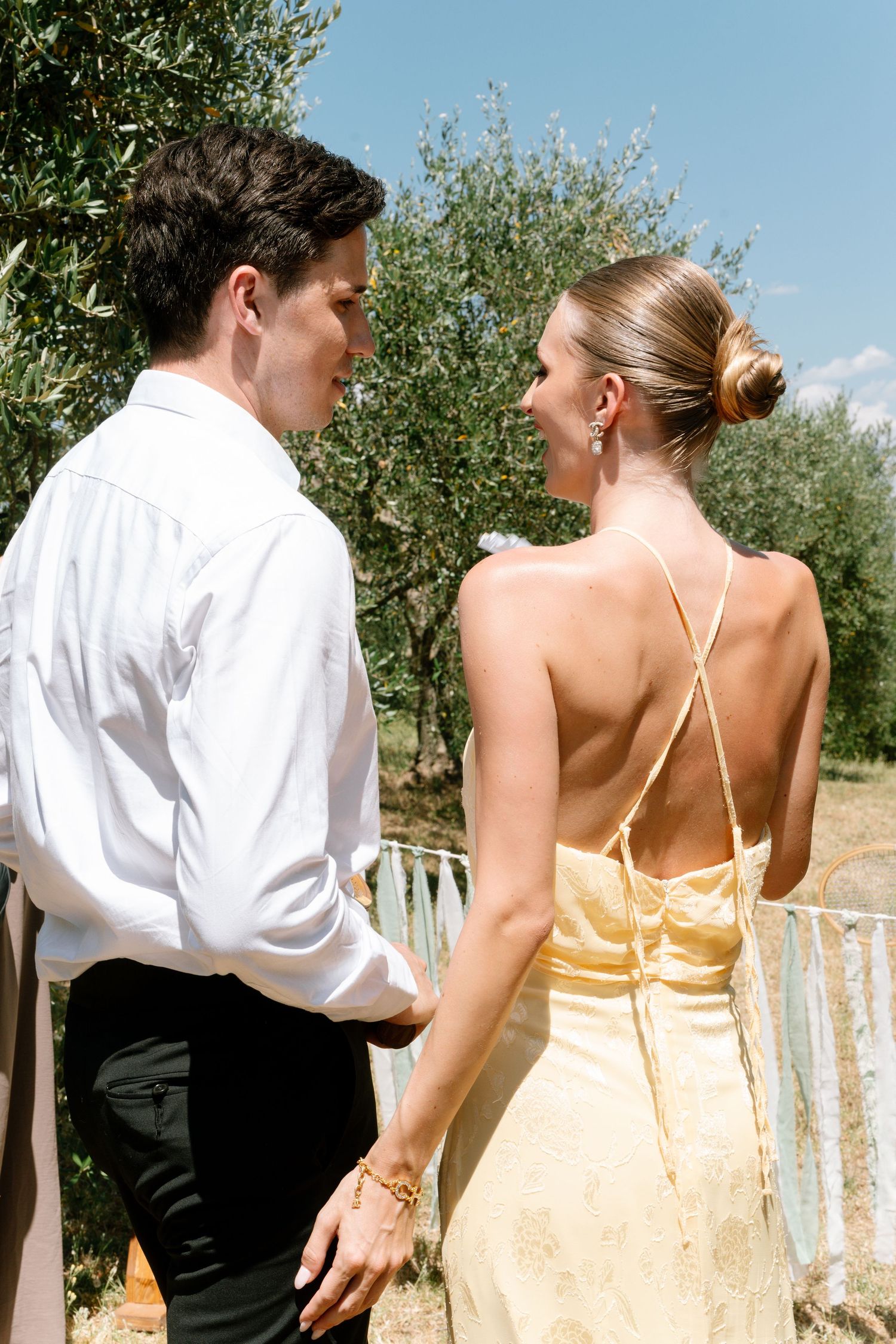 A couple in formal attire stands together in an olive grove on a sunny day.
