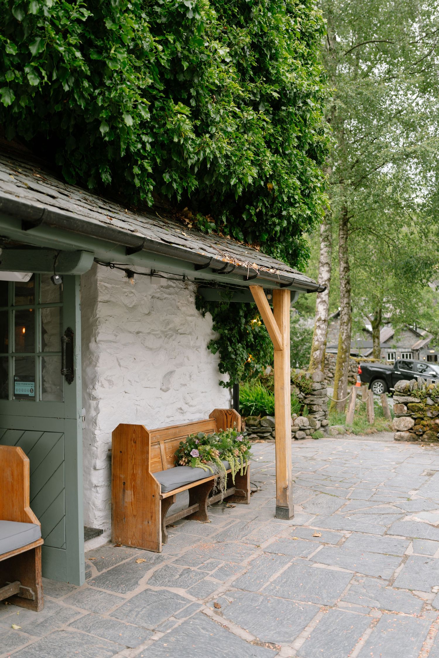 Stone patio with wooden benches and lush greenery against a whitewashed wall of a rustic building.