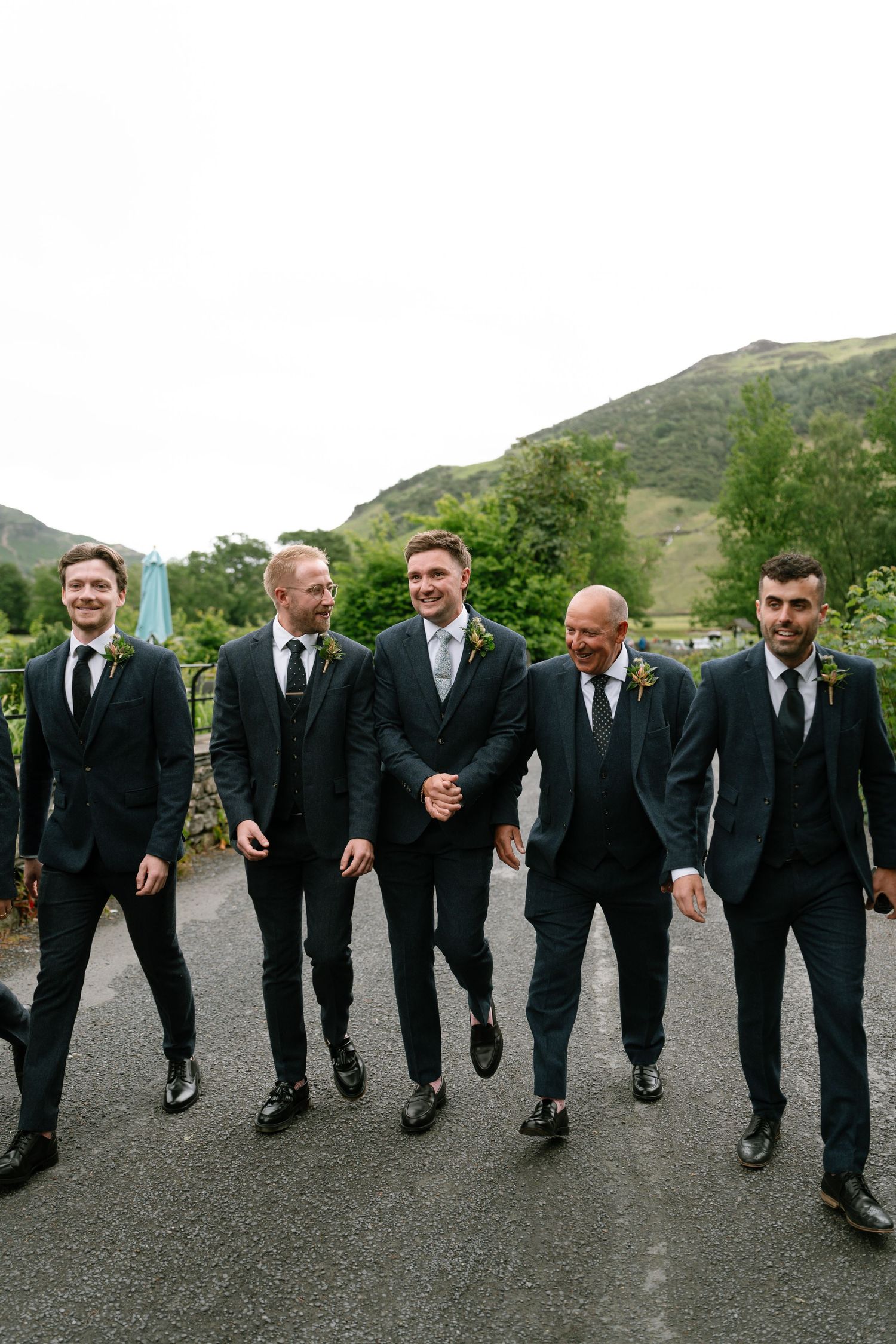 Groomsmen in black suits walking in line on outdoor path with mountains in background.