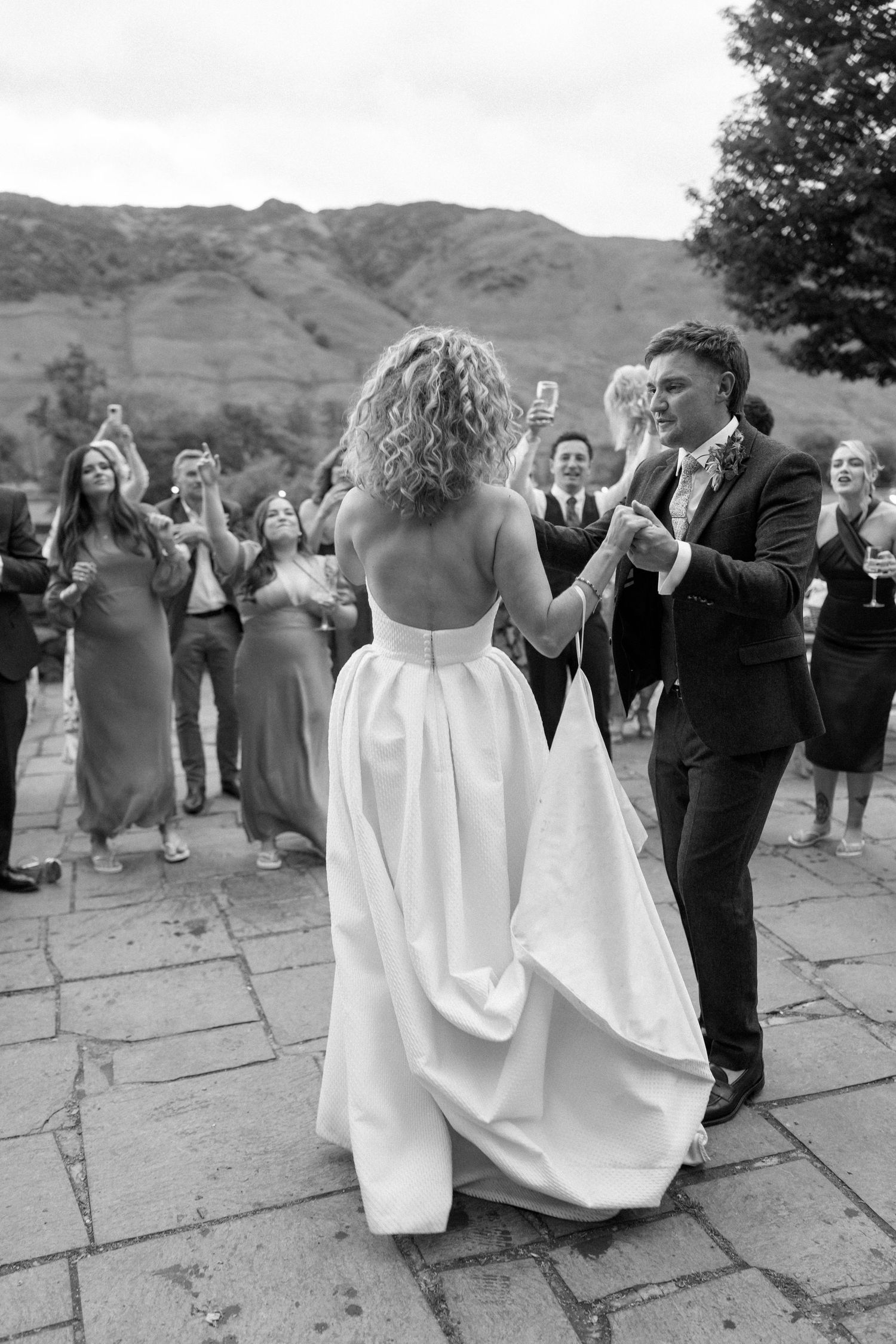 Black and white photo of wedding guests dancing outdoors with mountains visible in the distance.