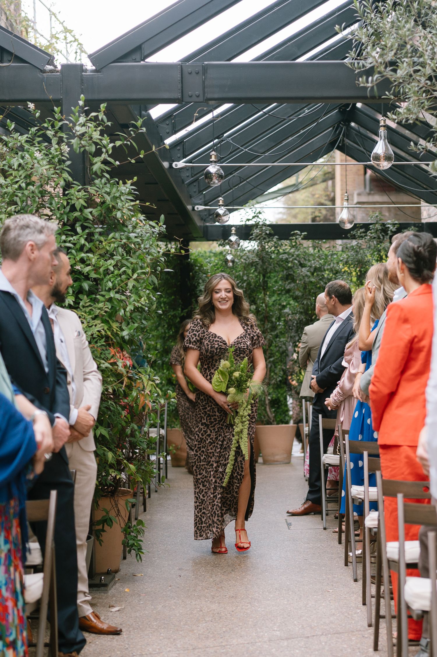 Wedding ceremony venue with glass ceiling and greenery, showing guests gathered in formal attire.