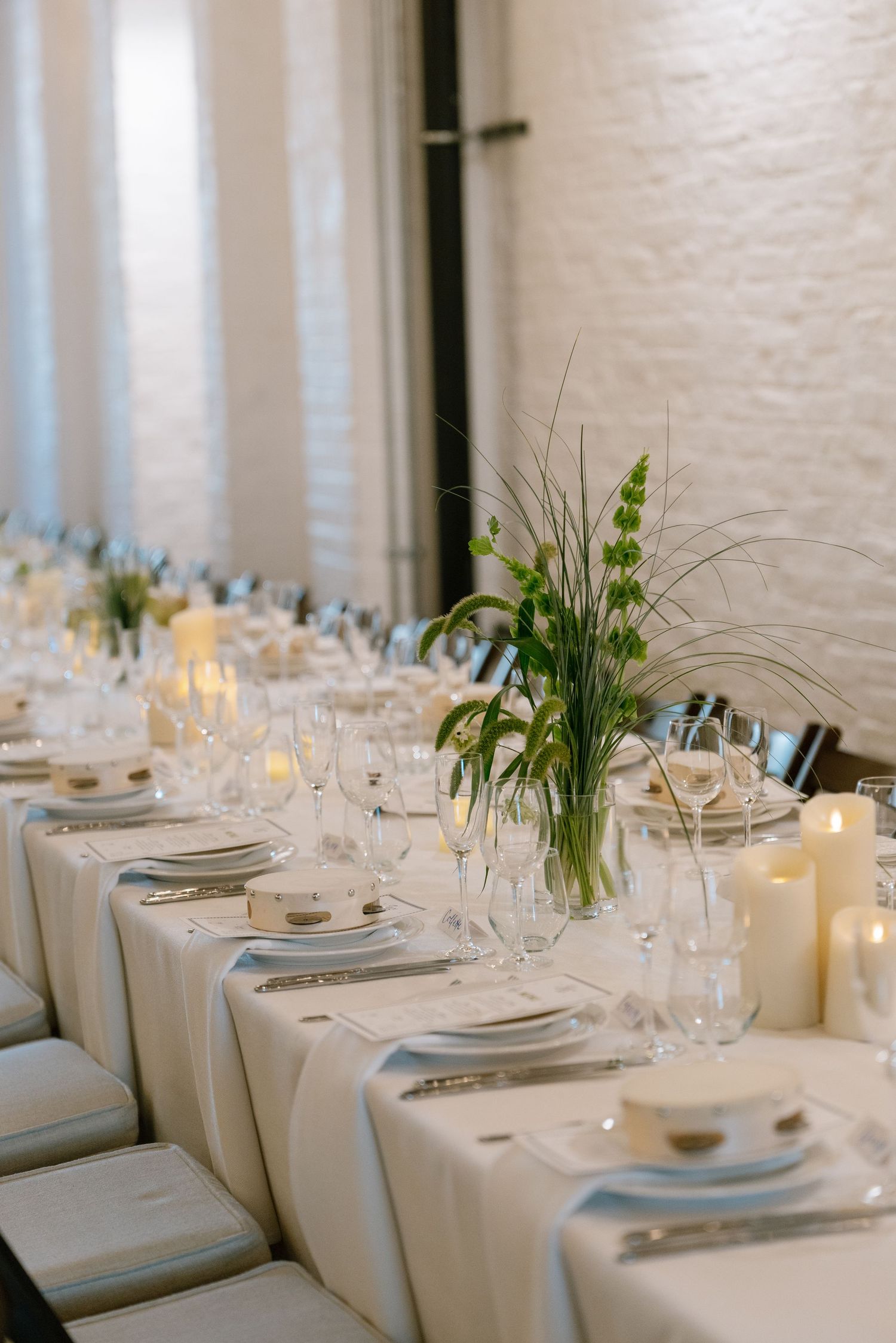 Modern minimalist wedding reception table setting with white linens, candles and simple greenery centerpieces.