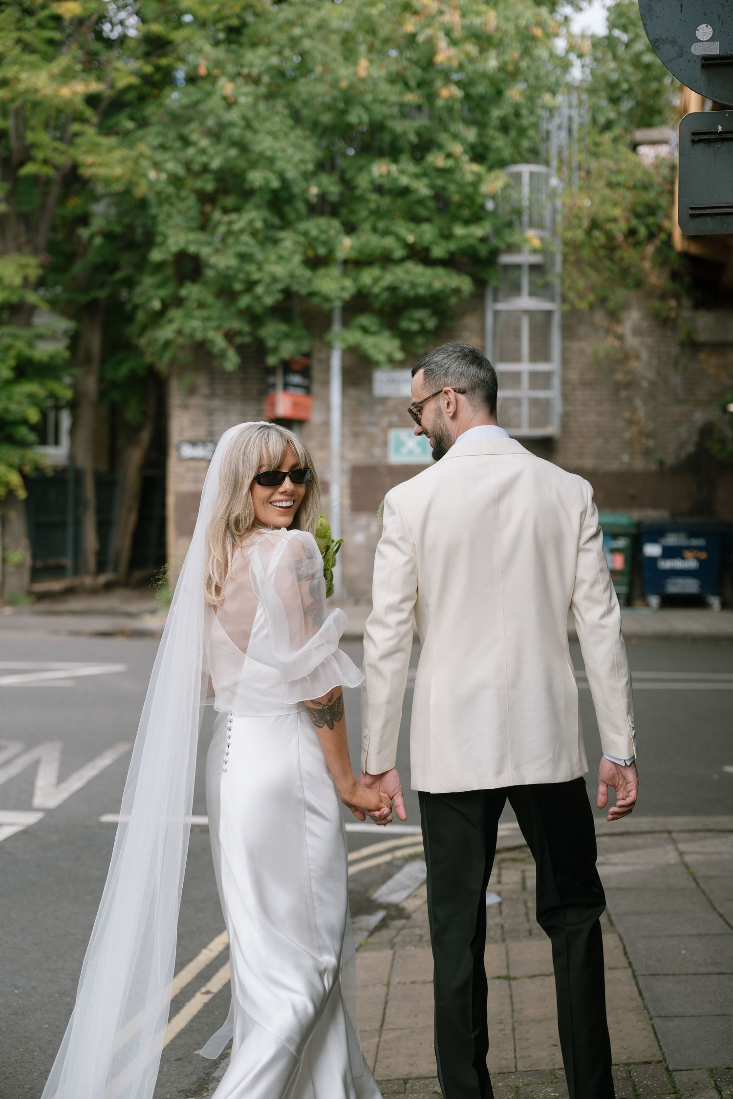 A couple in white formal attire walks hand in hand on an urban street.