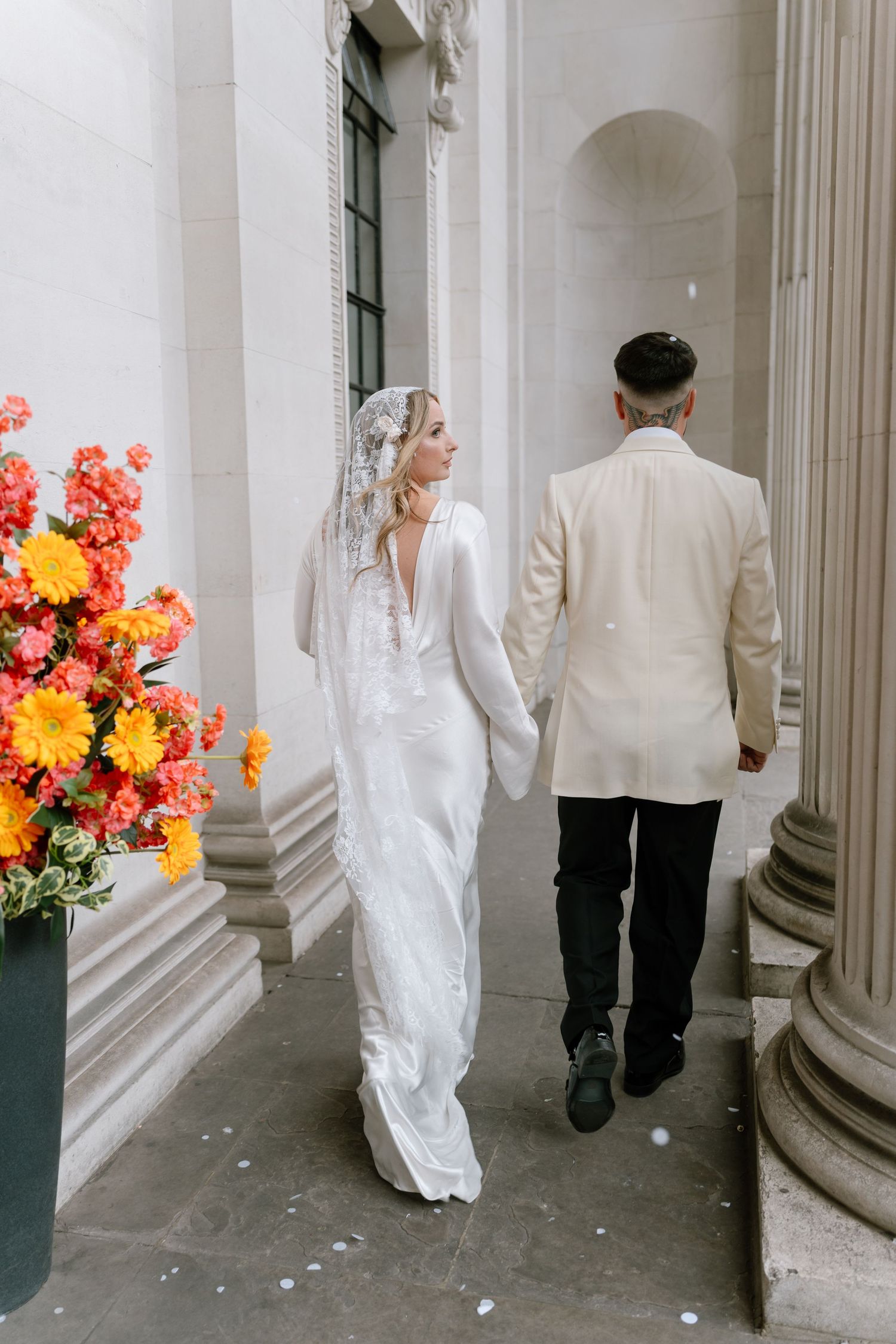 A wedding couple walks together through a columned entrance decorated with vibrant orange flowers.