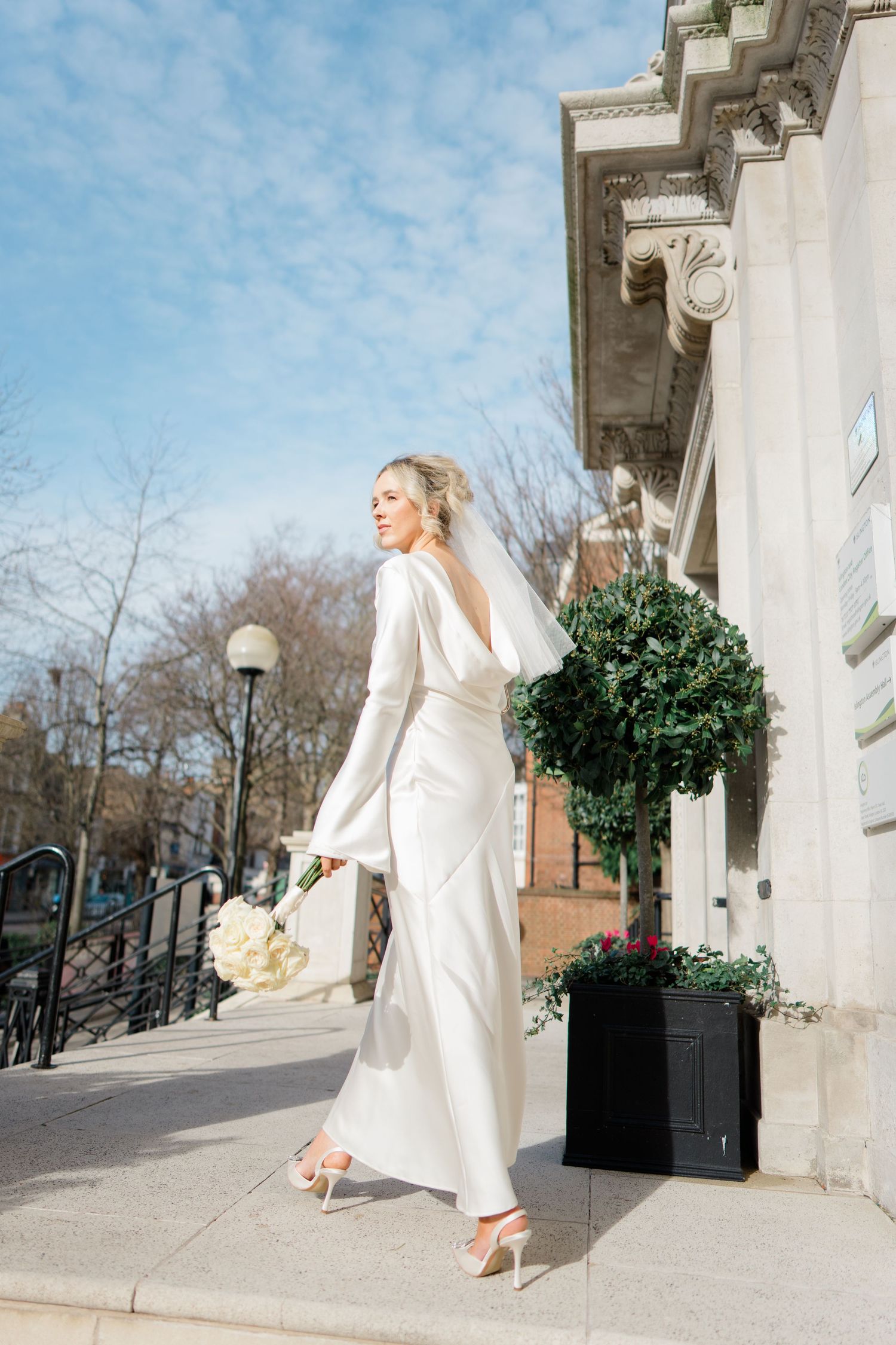 Bride in modern white pantsuit poses on classical building steps with decorative planters.
