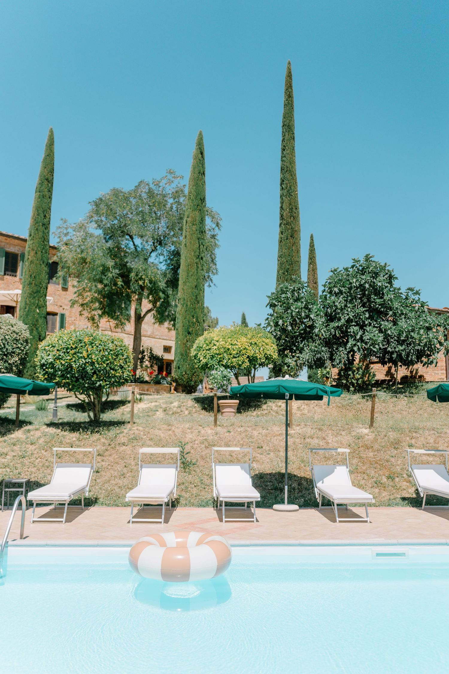 Poolside lounge chairs line up beside a swimming pool with cypress trees and a villa on a hillside in Tuscany.