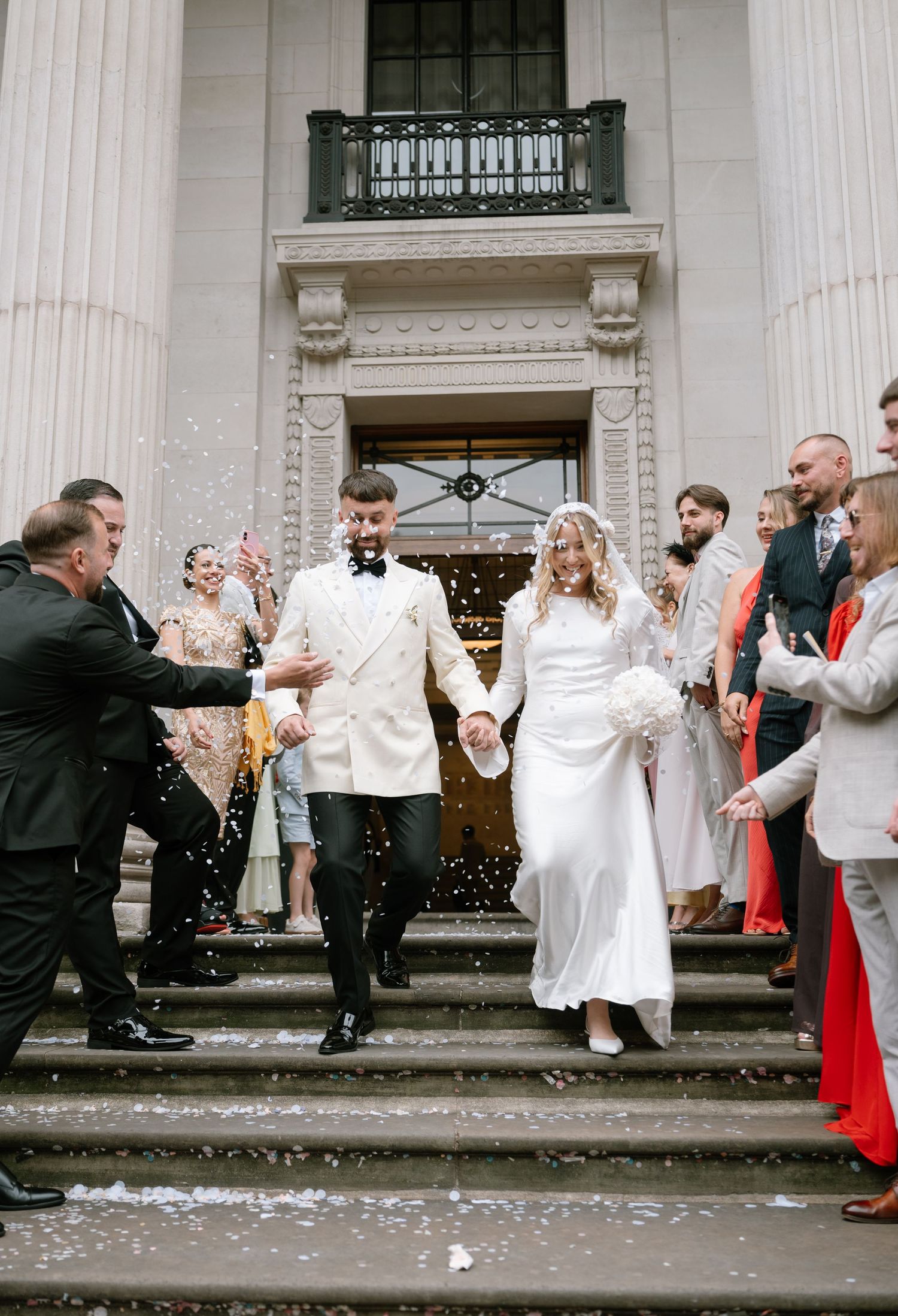 Newlyweds exit a grand building with white columns as guests shower them with confetti on the steps.