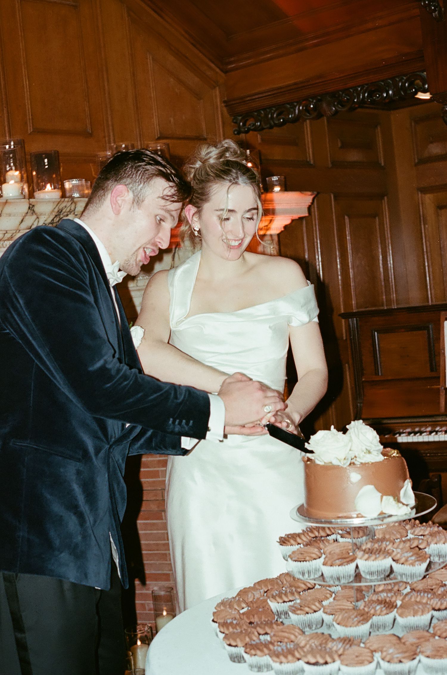 Couple cutting and sharing wedding cake surrounded by cupcakes at a wood-paneled reception.