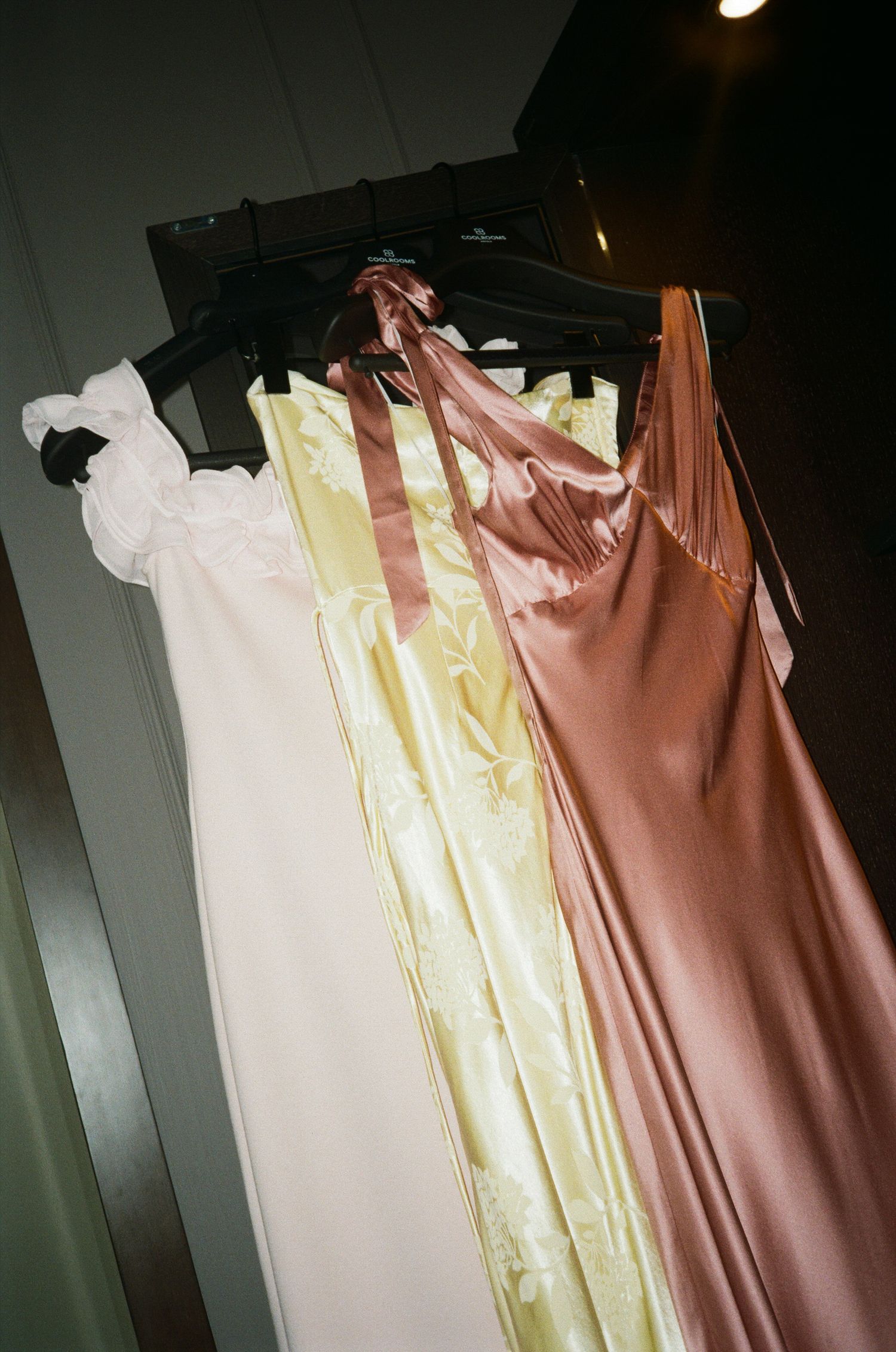 Three elegant dresses in white, cream and brown tones hang on display against a dark background.