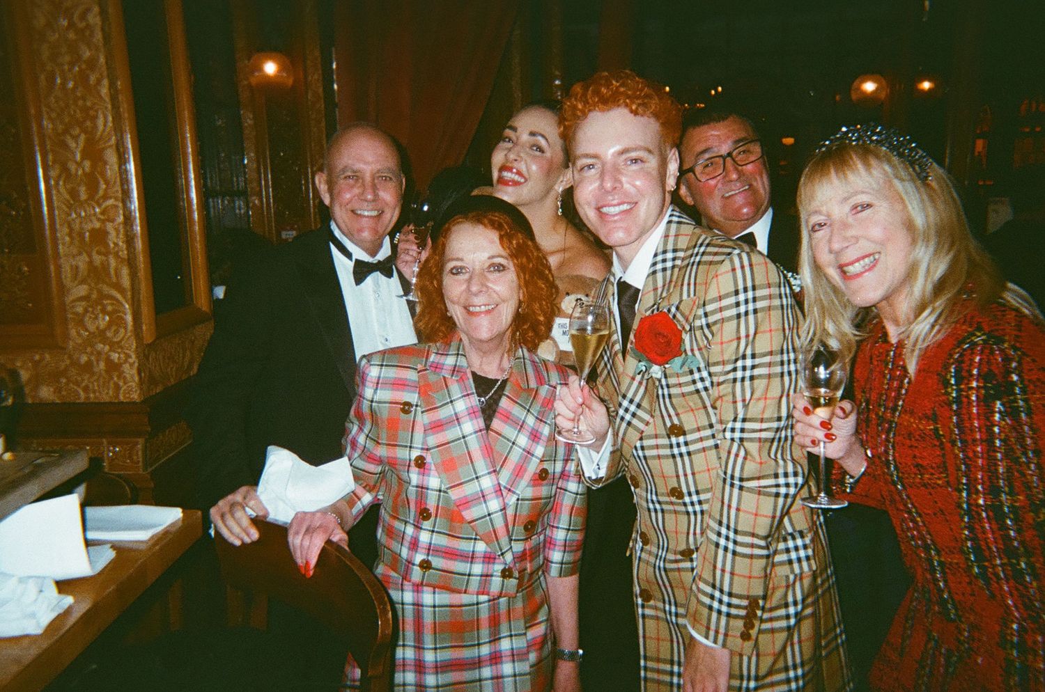 A group of people in formal and casual attire smile together at an evening social gathering.