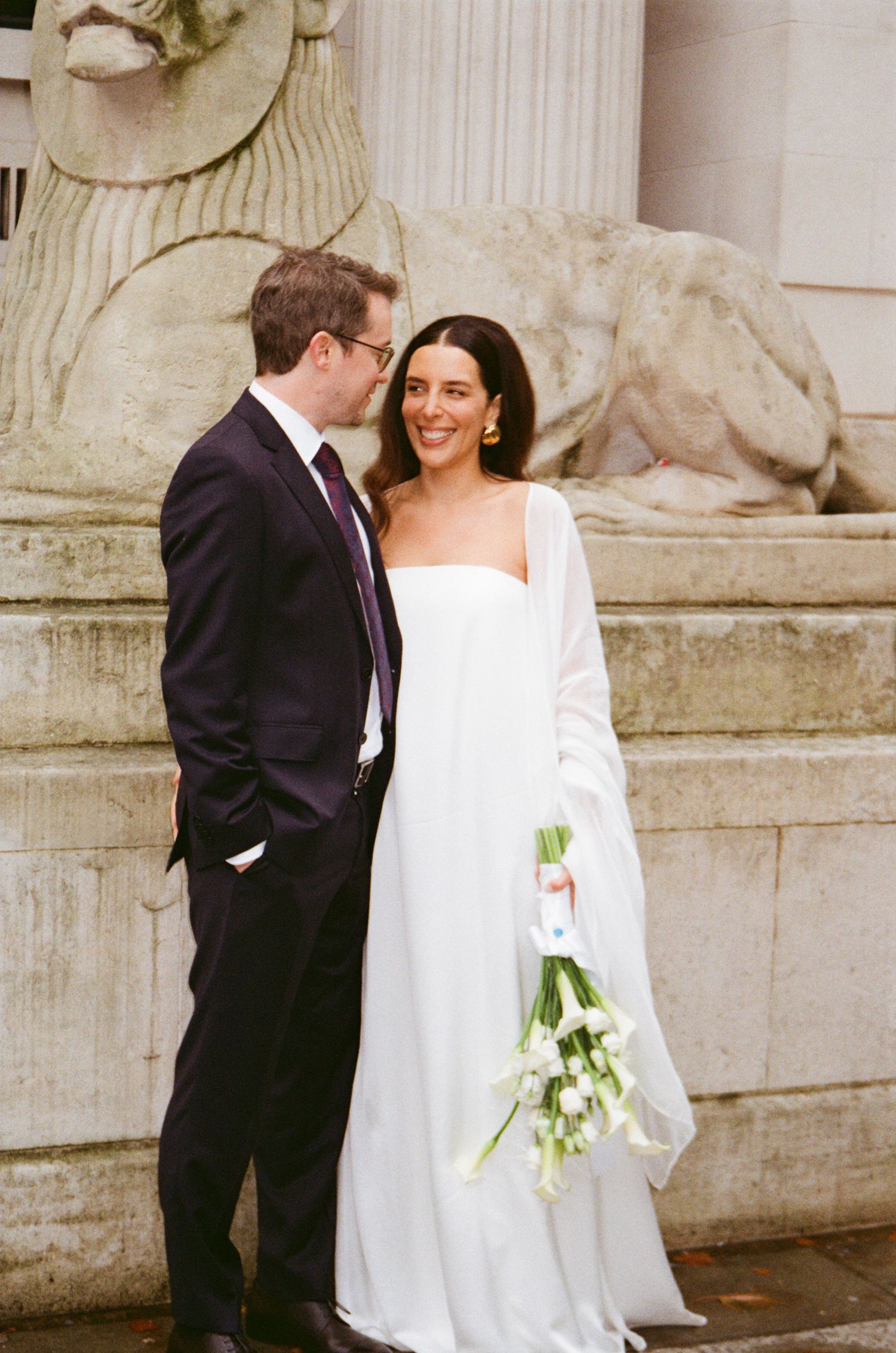 Bride in white long-sleeved dress holds bouquet while sharing intimate moment with groom by stone pillars.