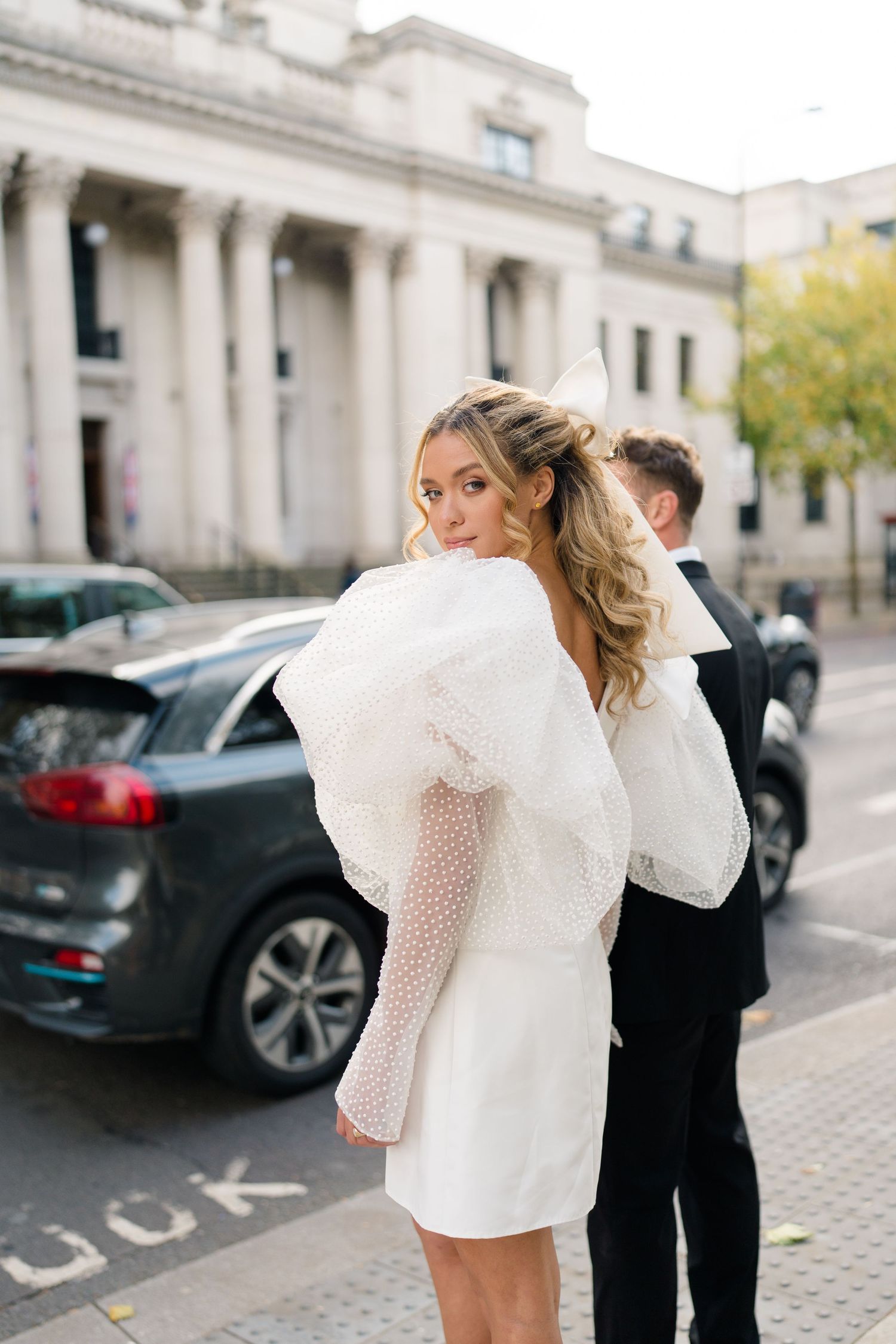 Couple in formal attire embracing outside classical building with luxury cars.