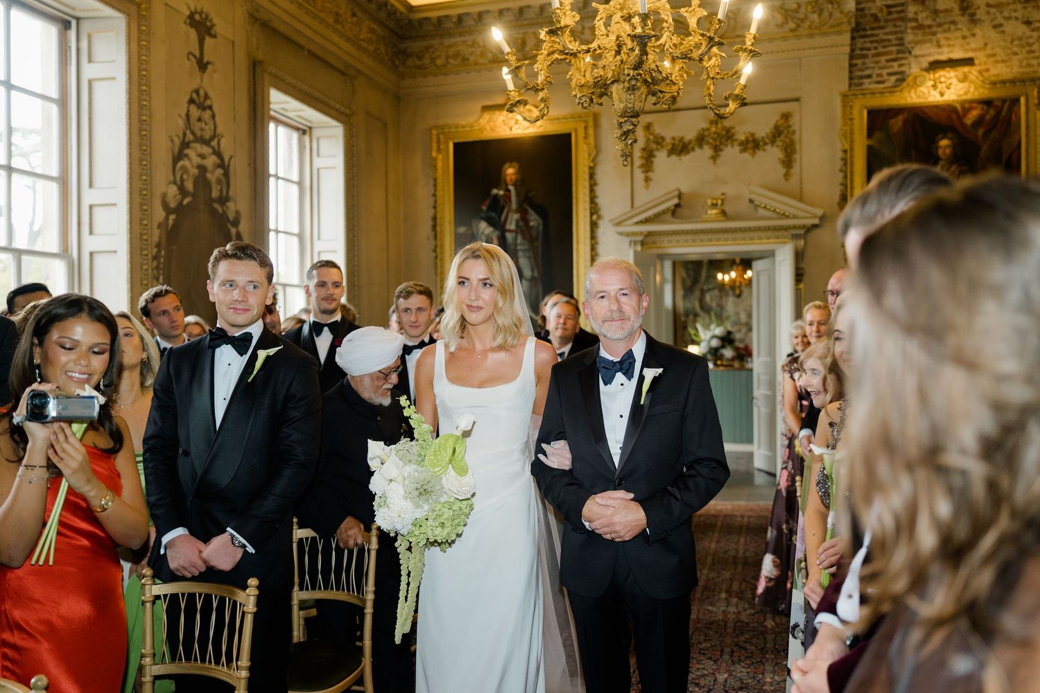 Bride in sleek white gown walking down the aisle with her father in a grand ornate ceremony room.