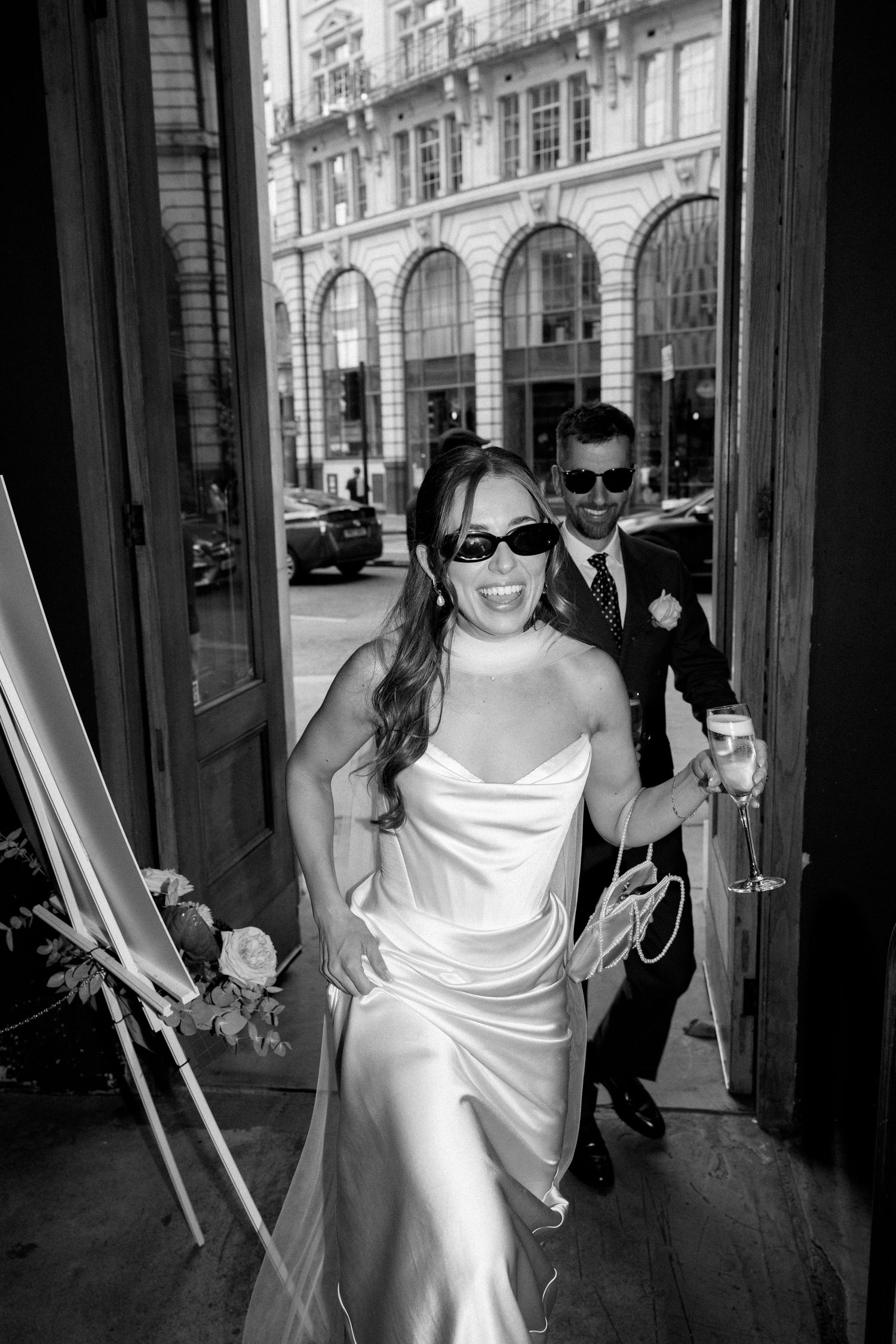 Bride in sleek white satin gown and sunglasses entering venue with groom in black and white photo.