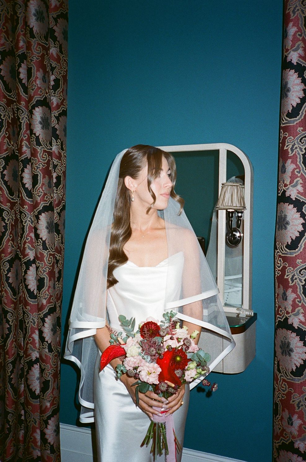 Beautiful bride in sleek white satin dress and veil holds red and pink bouquet beside vintage mirror in teal room.