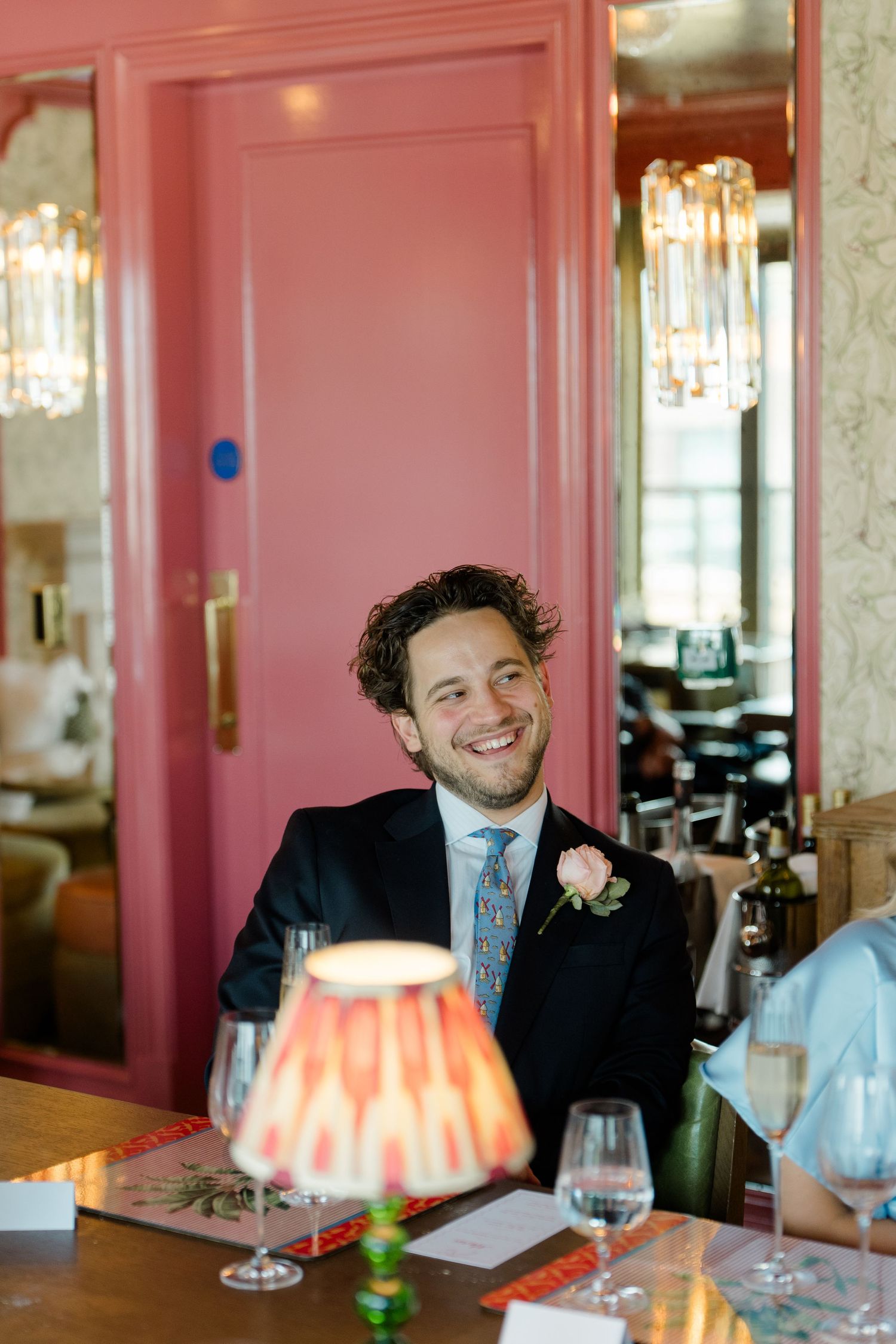 Smiling groom in suit with pink boutonniere seated at wedding reception table in elegant restaurant.