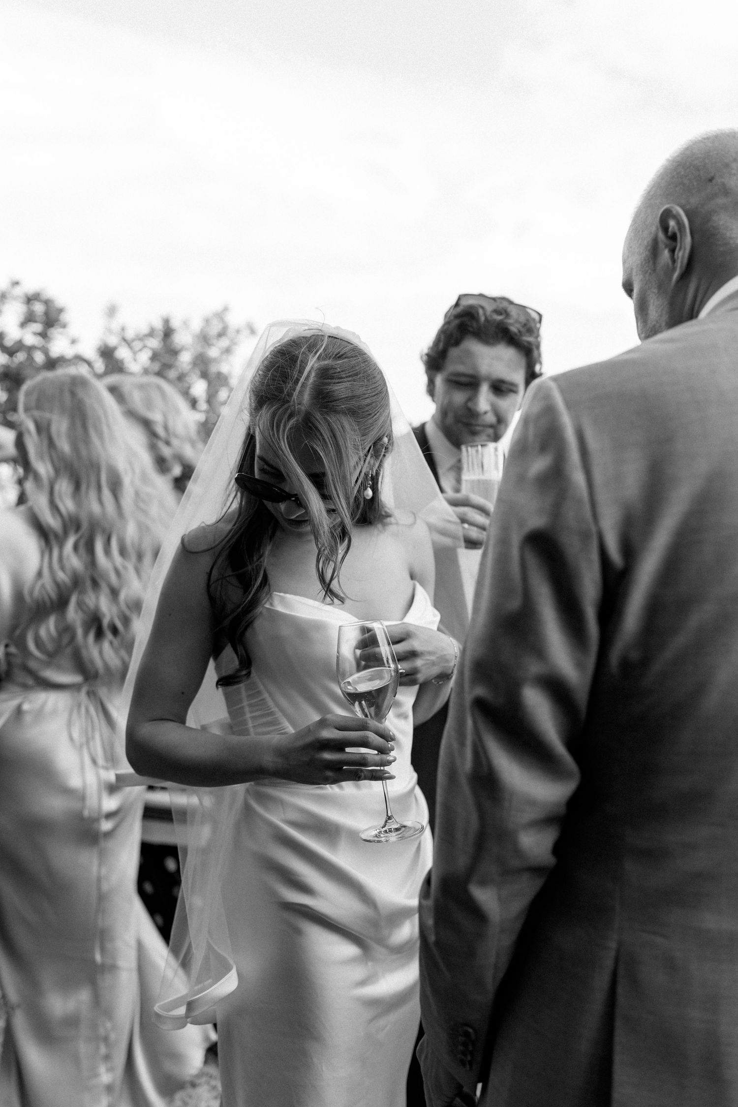 Bride in satin wedding dress holds champagne glass while chatting with guests at outdoor reception.