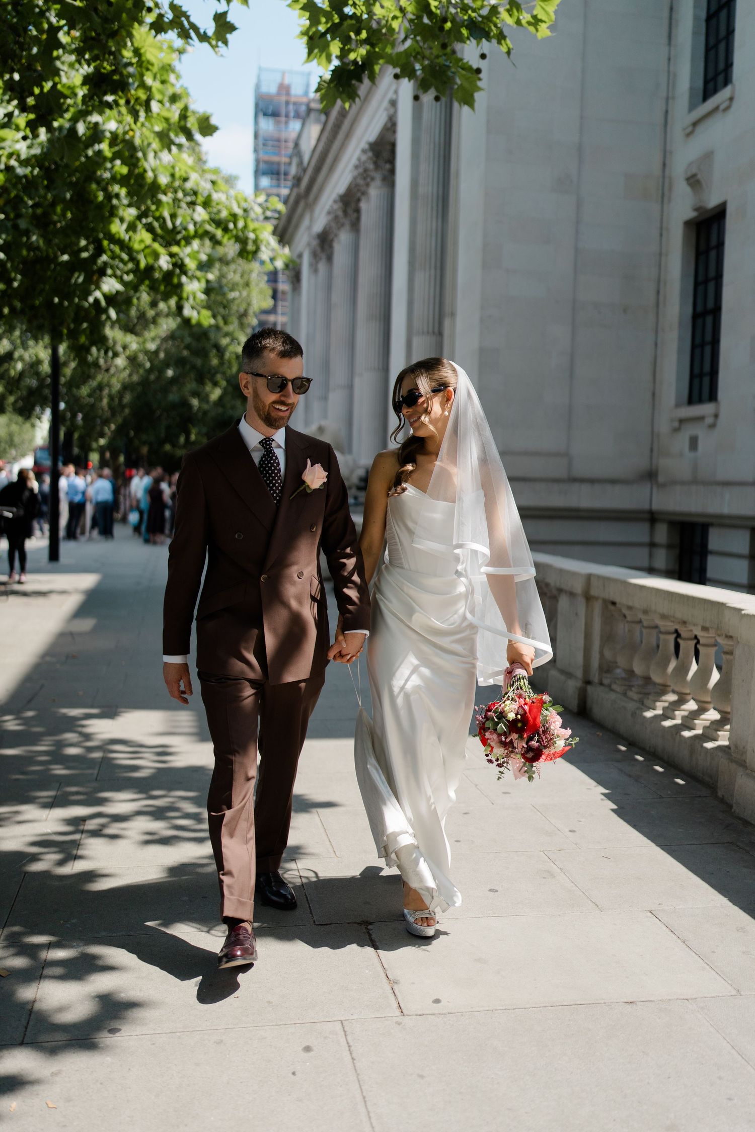 Stylish bride and groom walk hand-in-hand on sunny London street, wearing sunglasses and wedding attire.