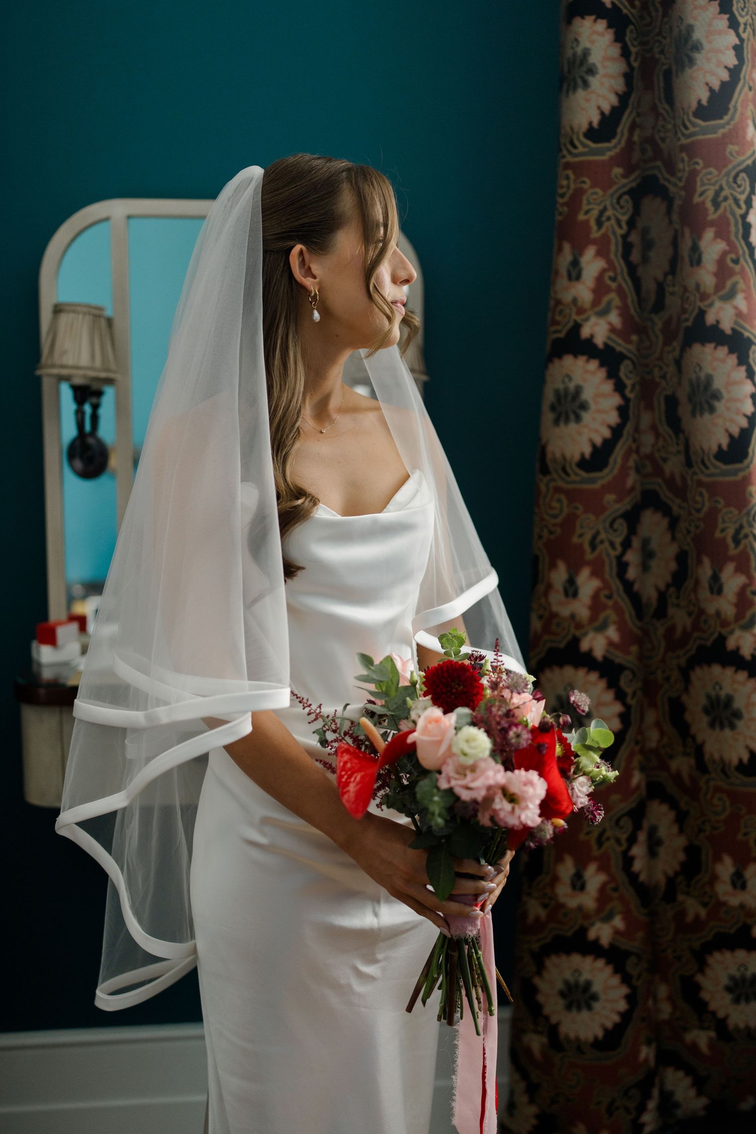 Bride in white satin gown and veil holds colorful bouquet of red and pink flowers indoors.