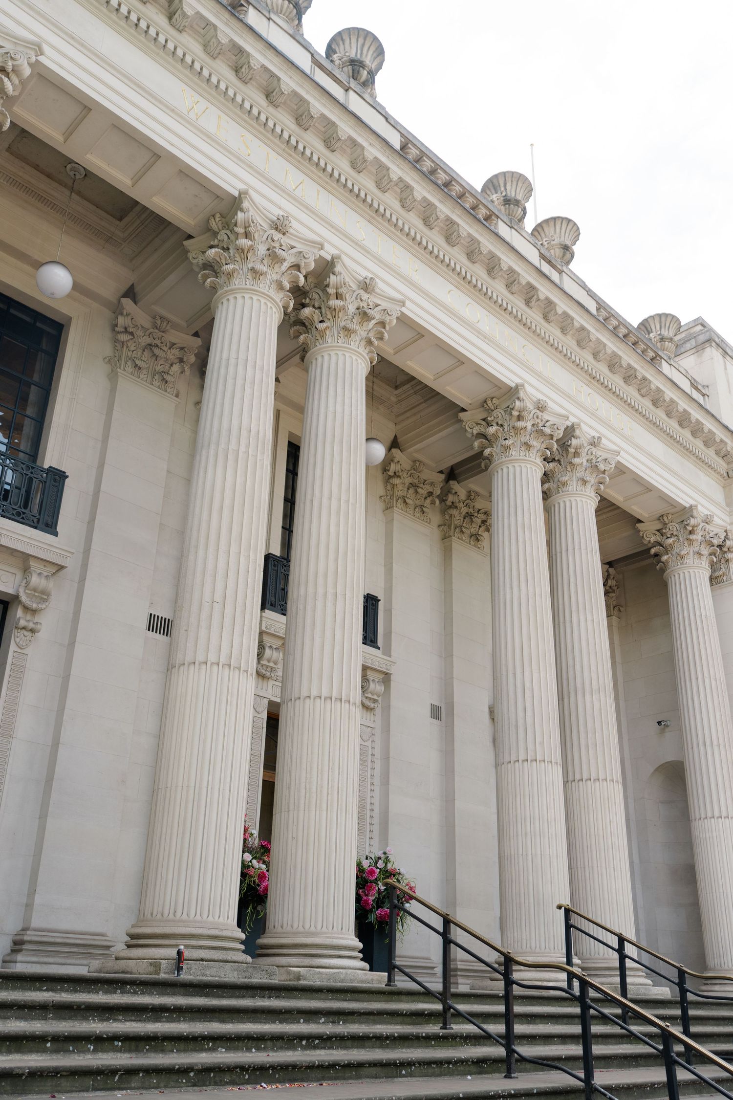 Grand neoclassical building exterior with tall Corinthian columns, stone steps, and floral arrangements at entrance.
