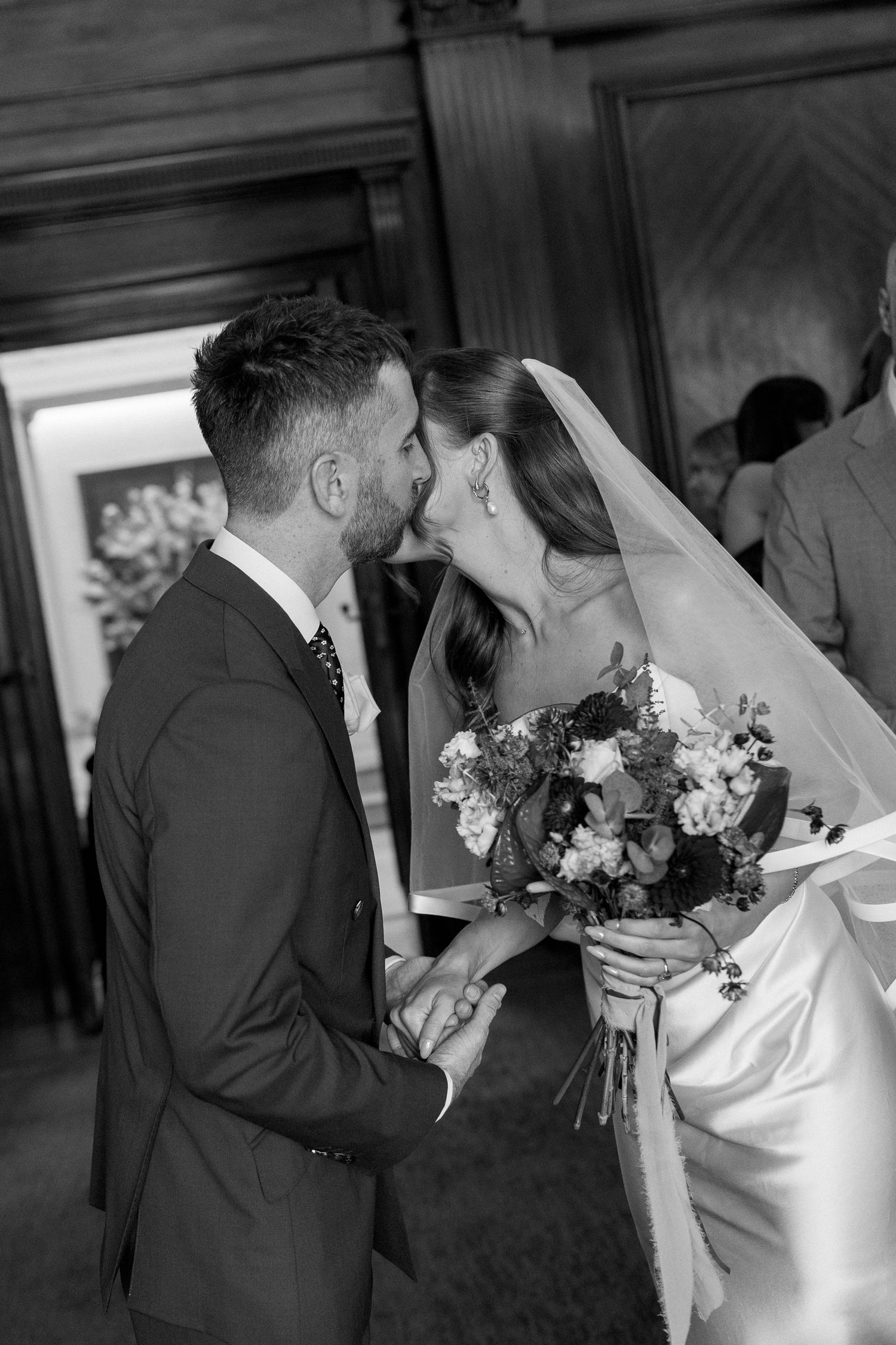 Black and white photo of newlyweds sharing first kiss during wedding ceremony with floral bouquet.