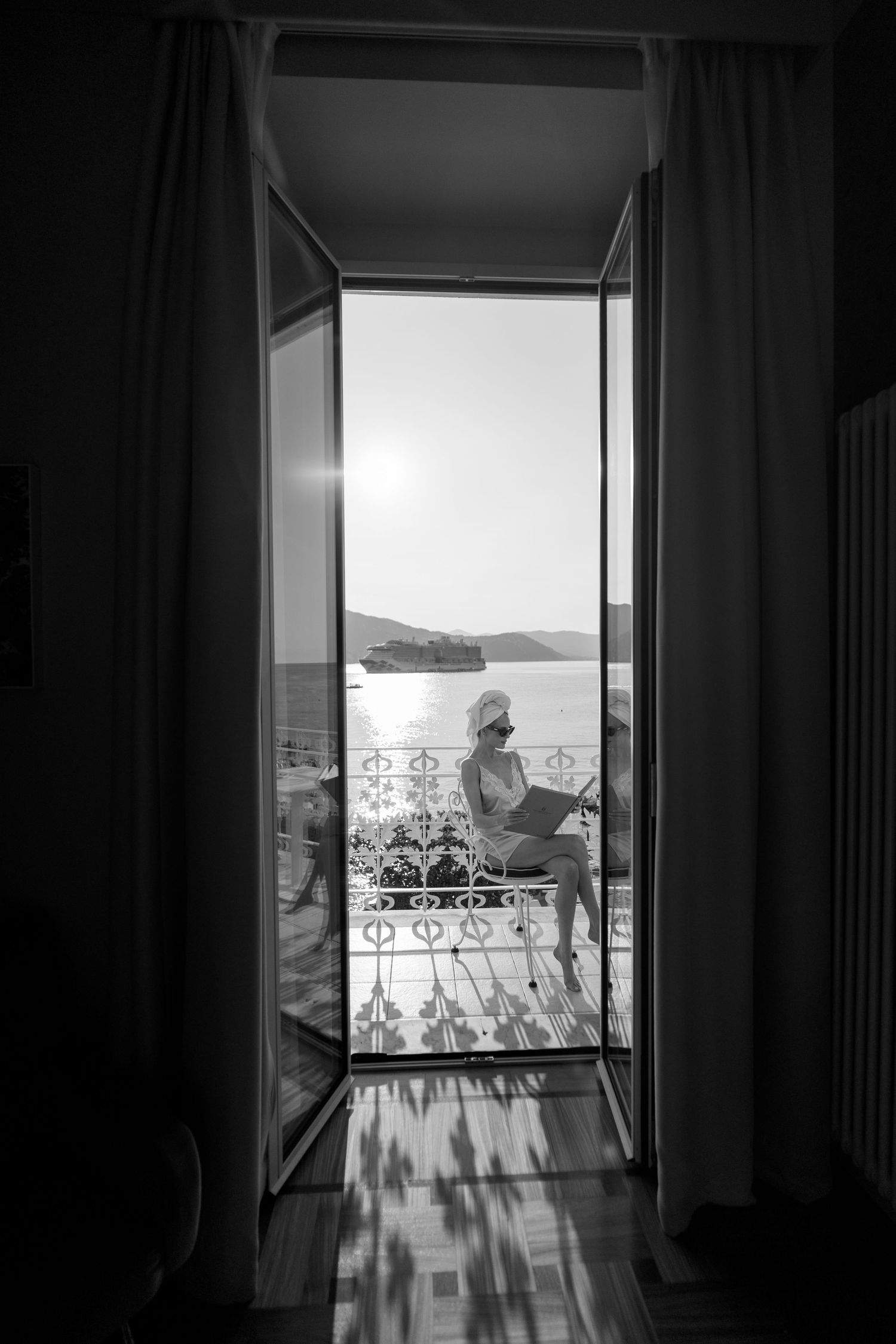 Woman in white towel reads on a sunlit balcony overlooking the sea, viewed through open French doors.