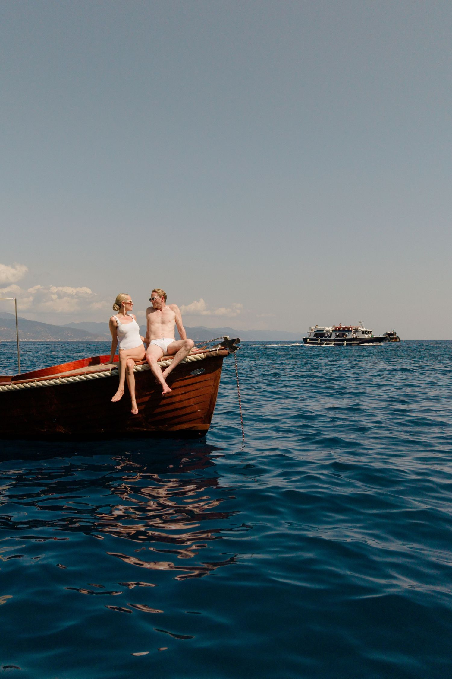 Couple in white swimwear sitting on the bow of a wooden boat on calm blue Mediterranean sea waters.
