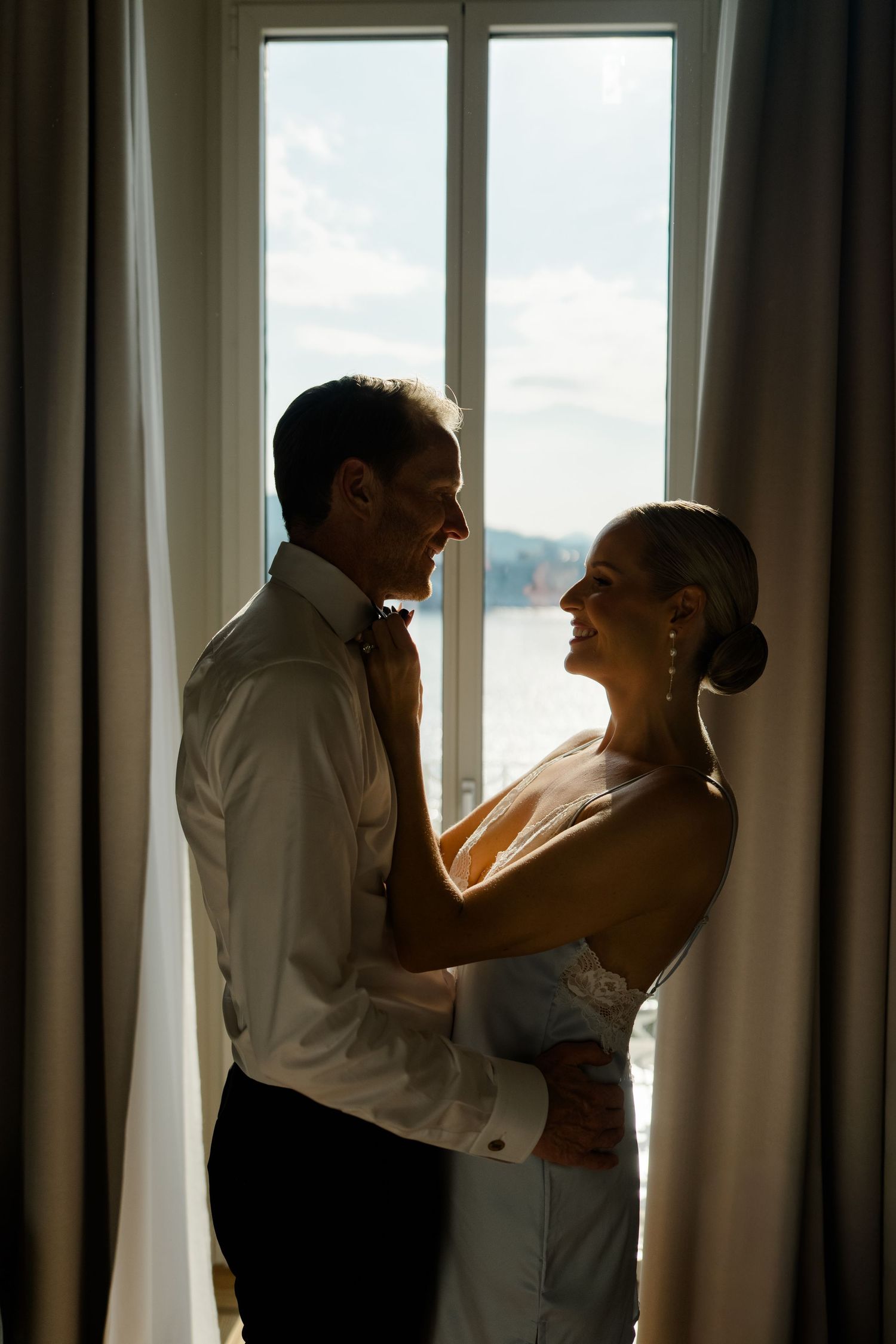 Bride and groom share intimate moment silhouetted against large window with ocean view, smiling at each other.