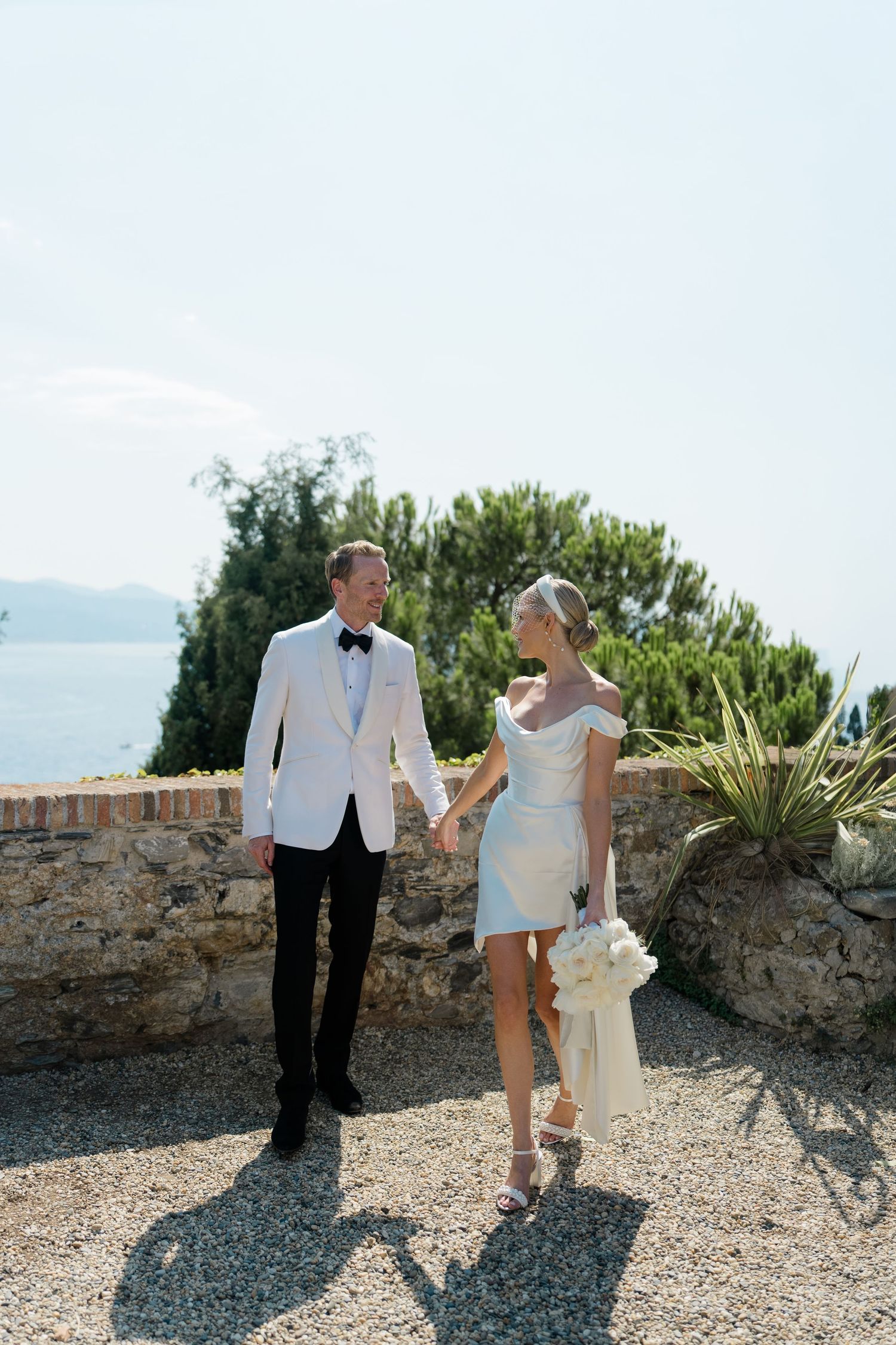 Elegant bride and groom holding hands walking along a stone terrace with scenic Mediterranean sea views.