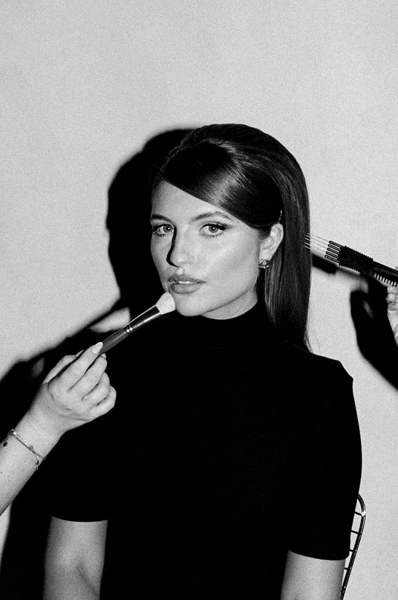 Woman in black top getting makeup applied with brushes in black and white photo.