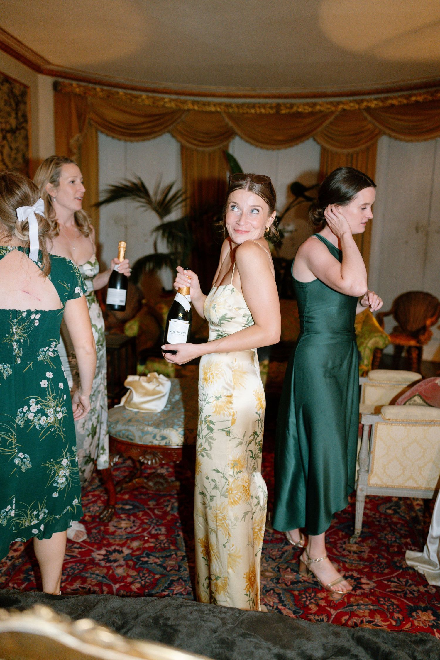 Wedding party members in green dresses and floral gowns hold champagne bottles during celebration.