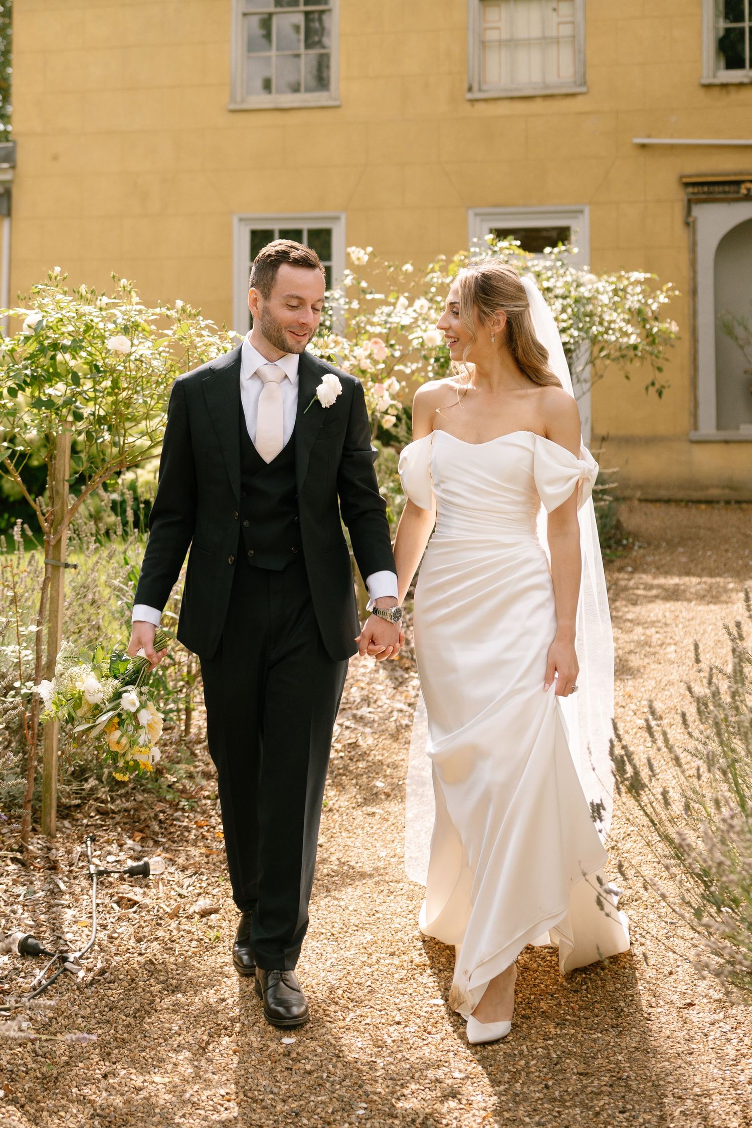 A couple in wedding attire strolls along a gravel garden path beside a yellow stone building.